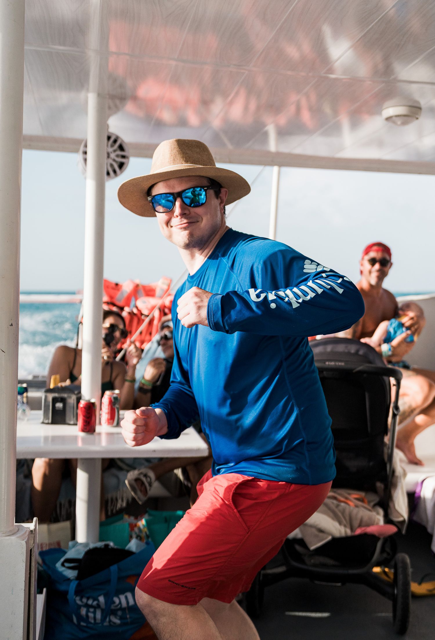 Man in blue Columbia shirt, red shorts, and straw hat dancing on a boat with people in background.