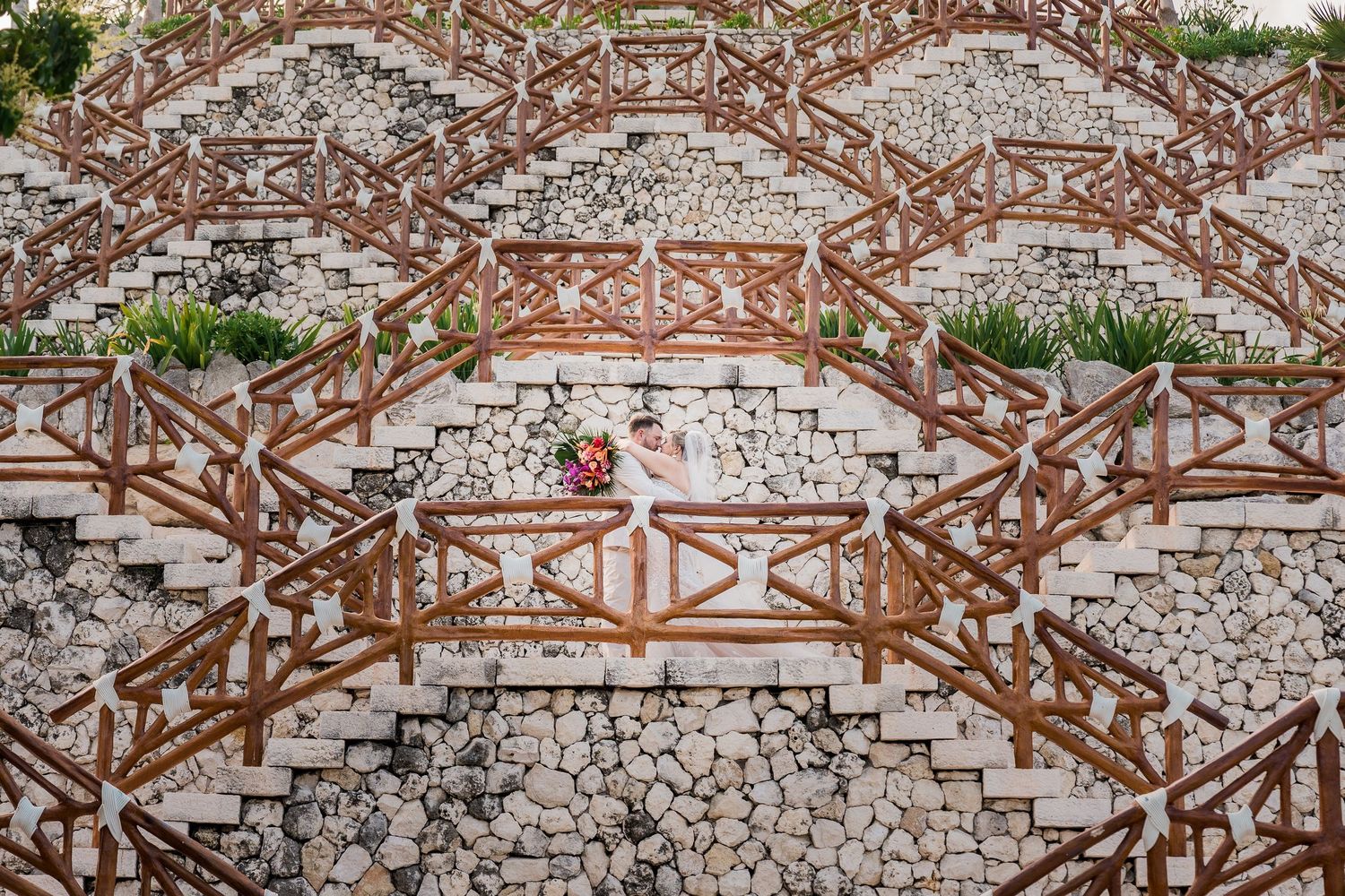 Aerial view of a bride in white dress posing on ornate stone staircase with rustic wooden railings.