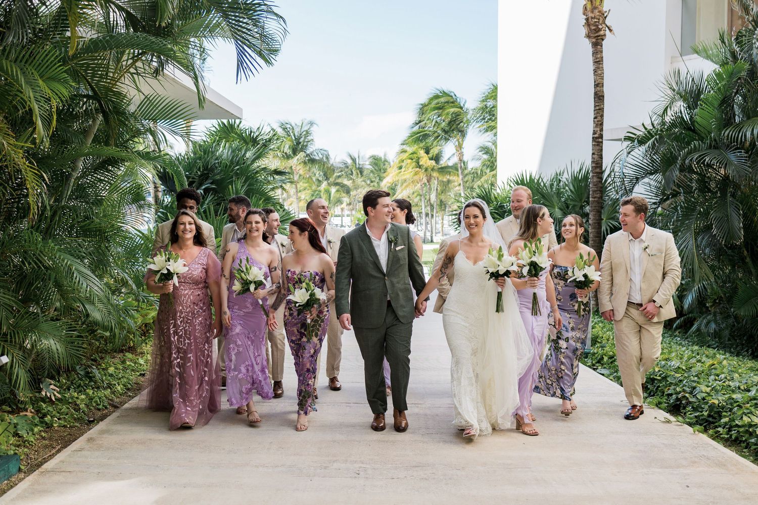 Wedding party walks together outdoors at tropical resort, bride and groom centered with bridesmaids in purple dresses and groomsmen.