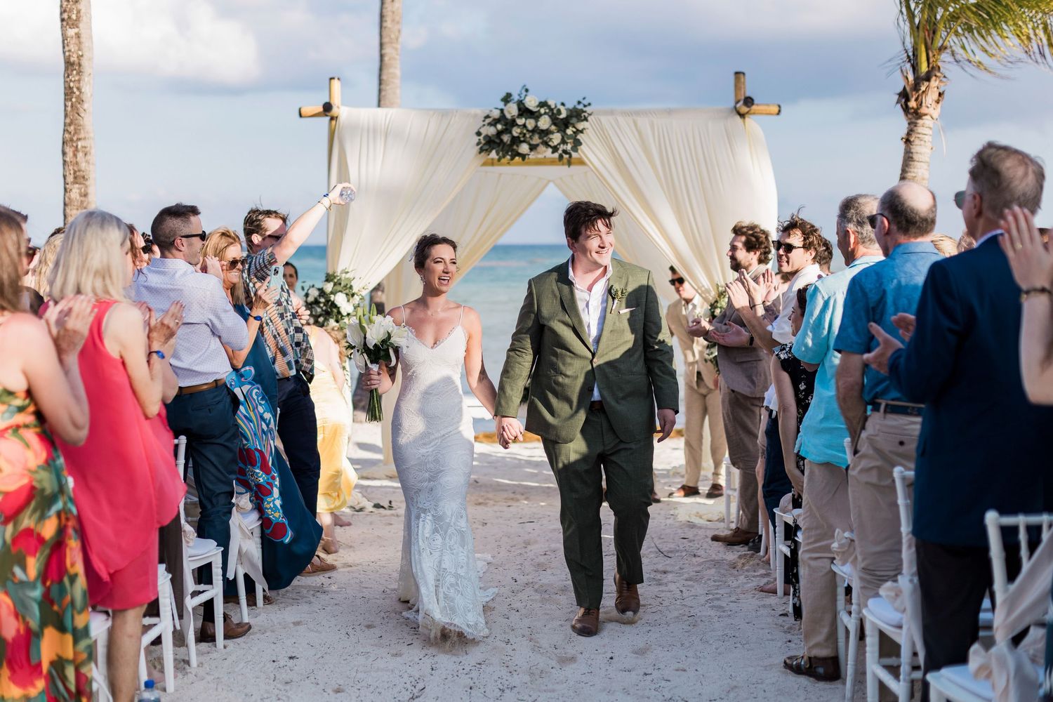 Newlywed couple walks down beach aisle hand-in-hand under floral arch, surrounded by cheering guests at outdoor wedding.