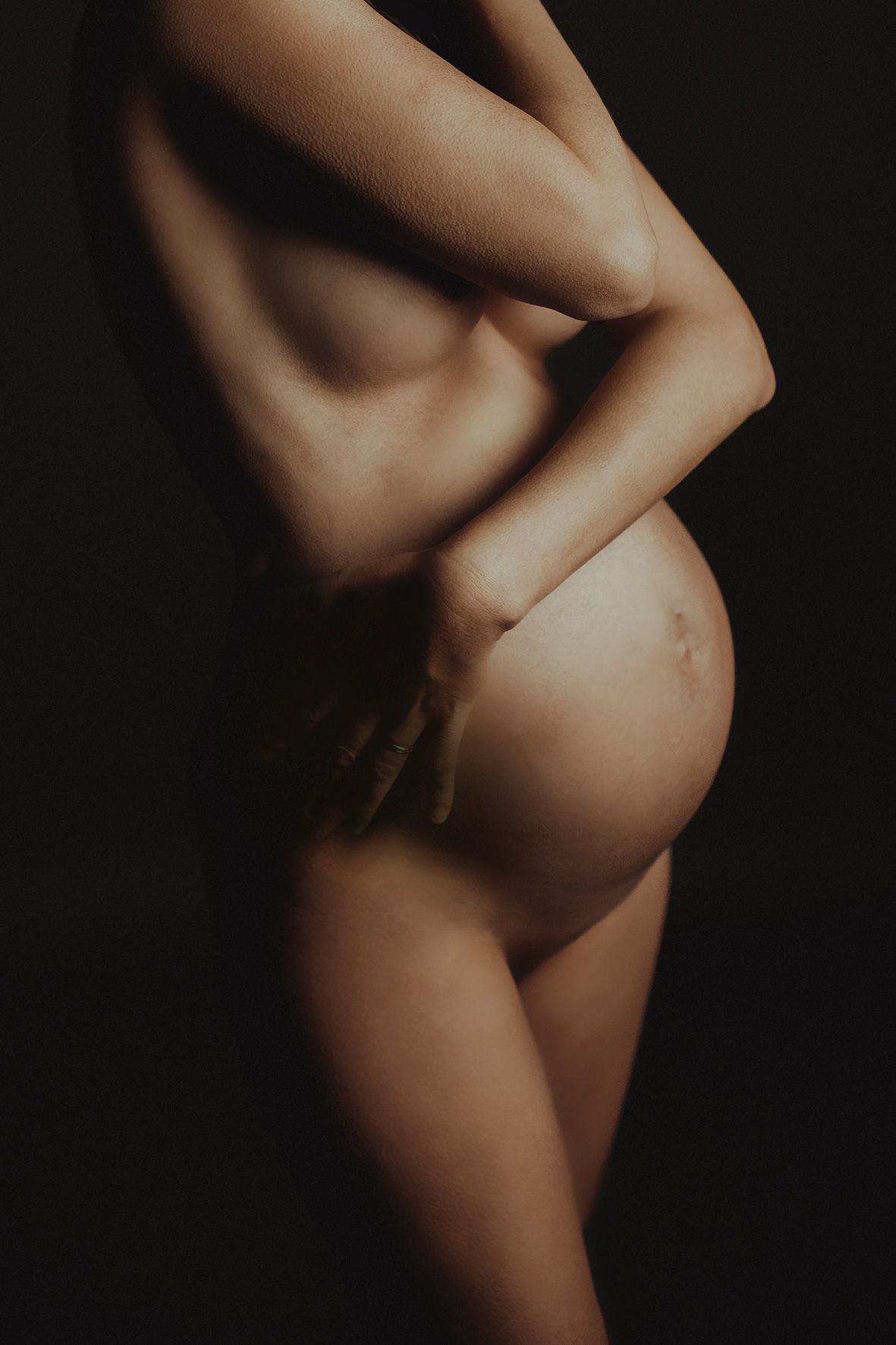 Artistic nude silhouette photograph of a pregnant body against dark background showcasing the natural curves of maternity.