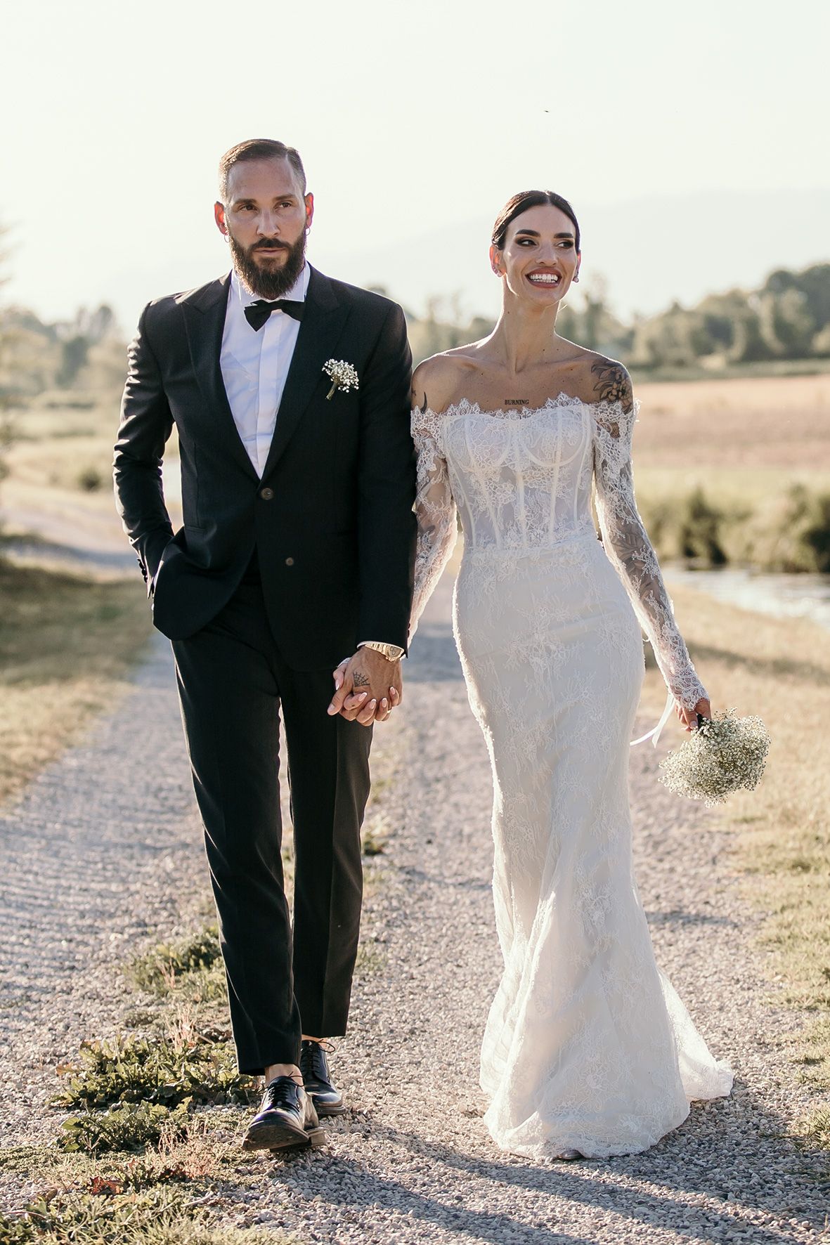 A couple walks hand in hand on a dirt path, with the bride in a lace white gown and groom in classic black tuxedo.