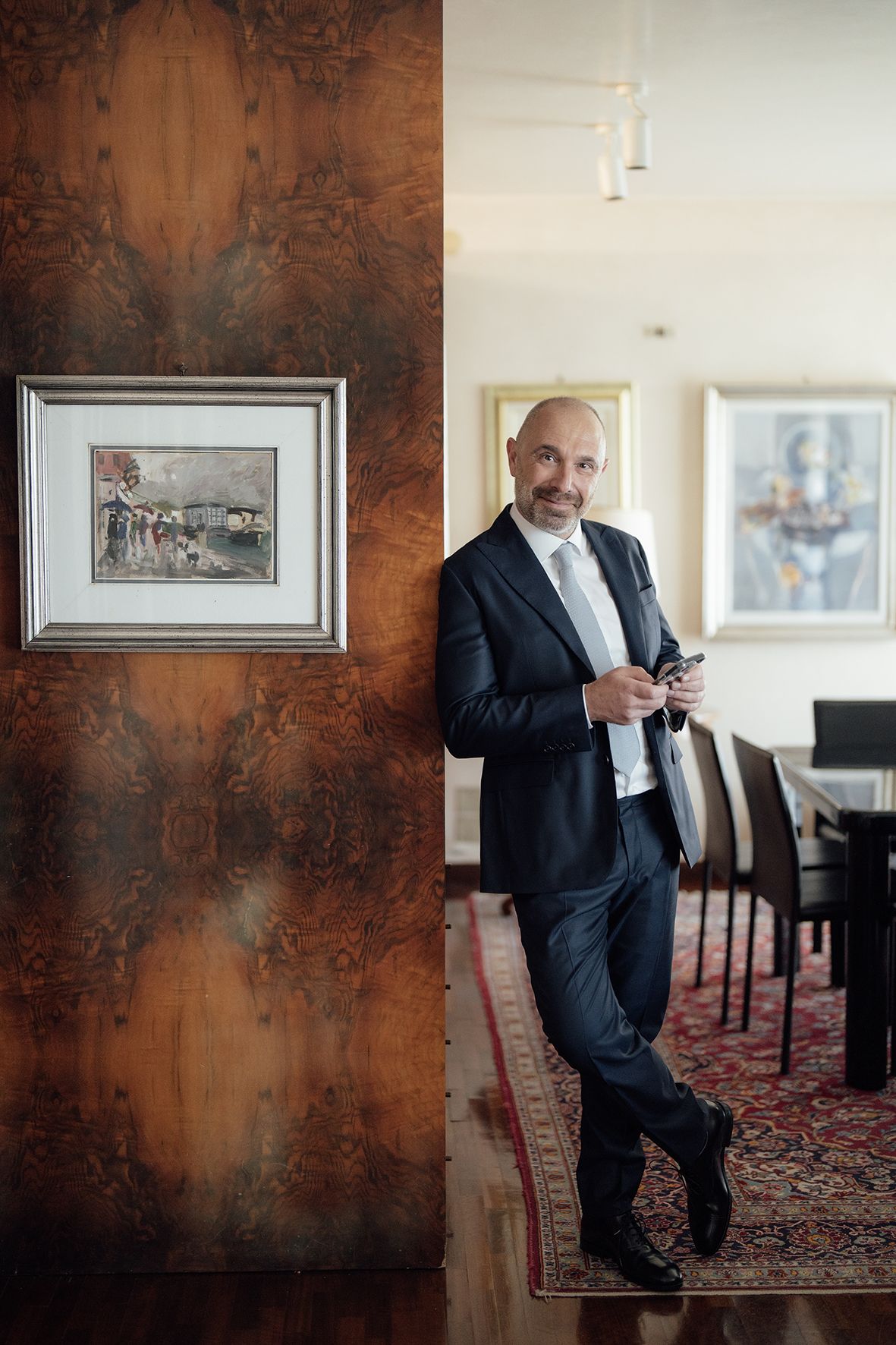 Professional in dark suit leans against textured wall near framed artwork in elegant interior setting.