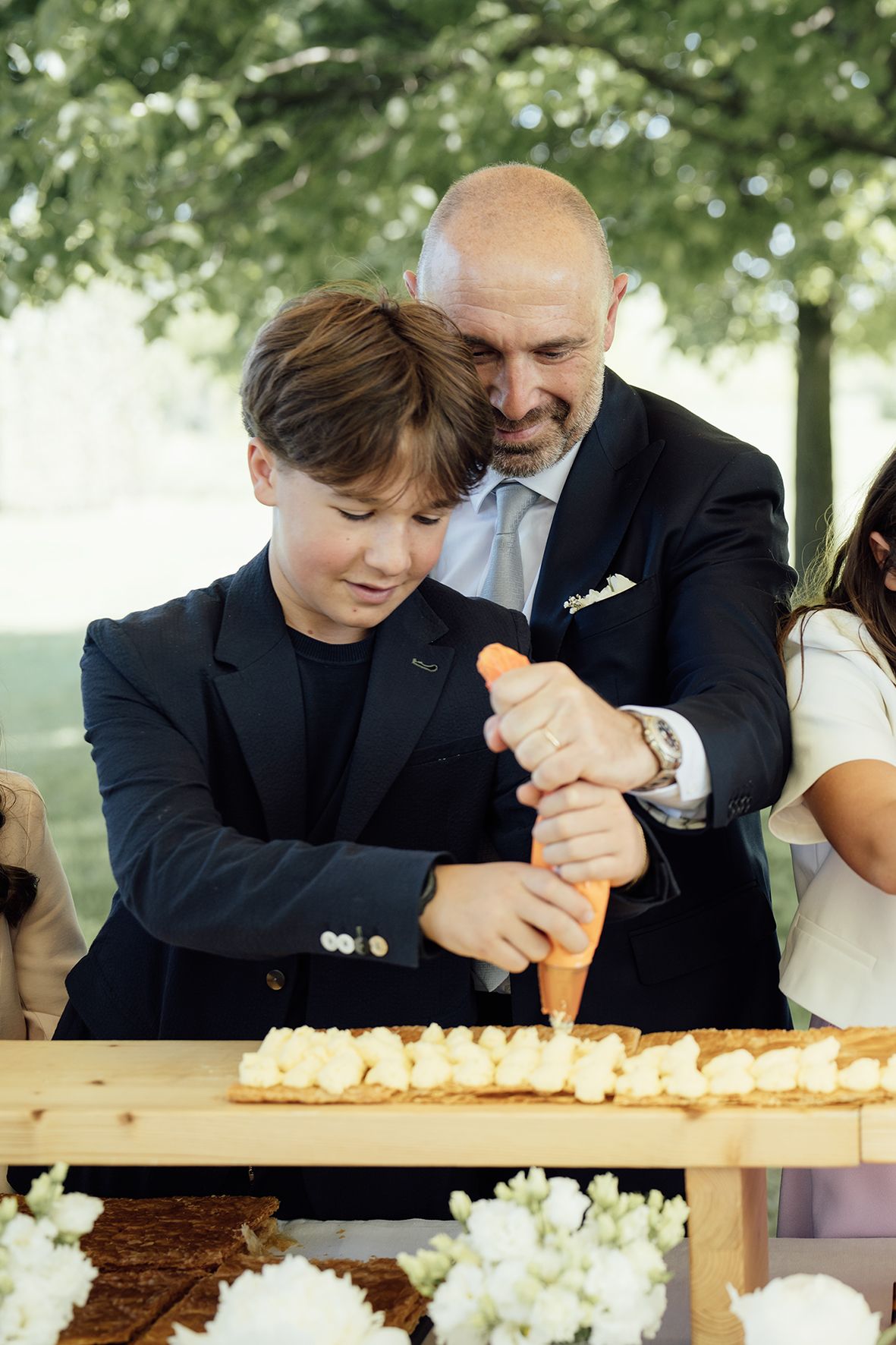 Someone in a dark suit squeezes icing onto a dessert table while being assisted at an elegant outdoor event.
