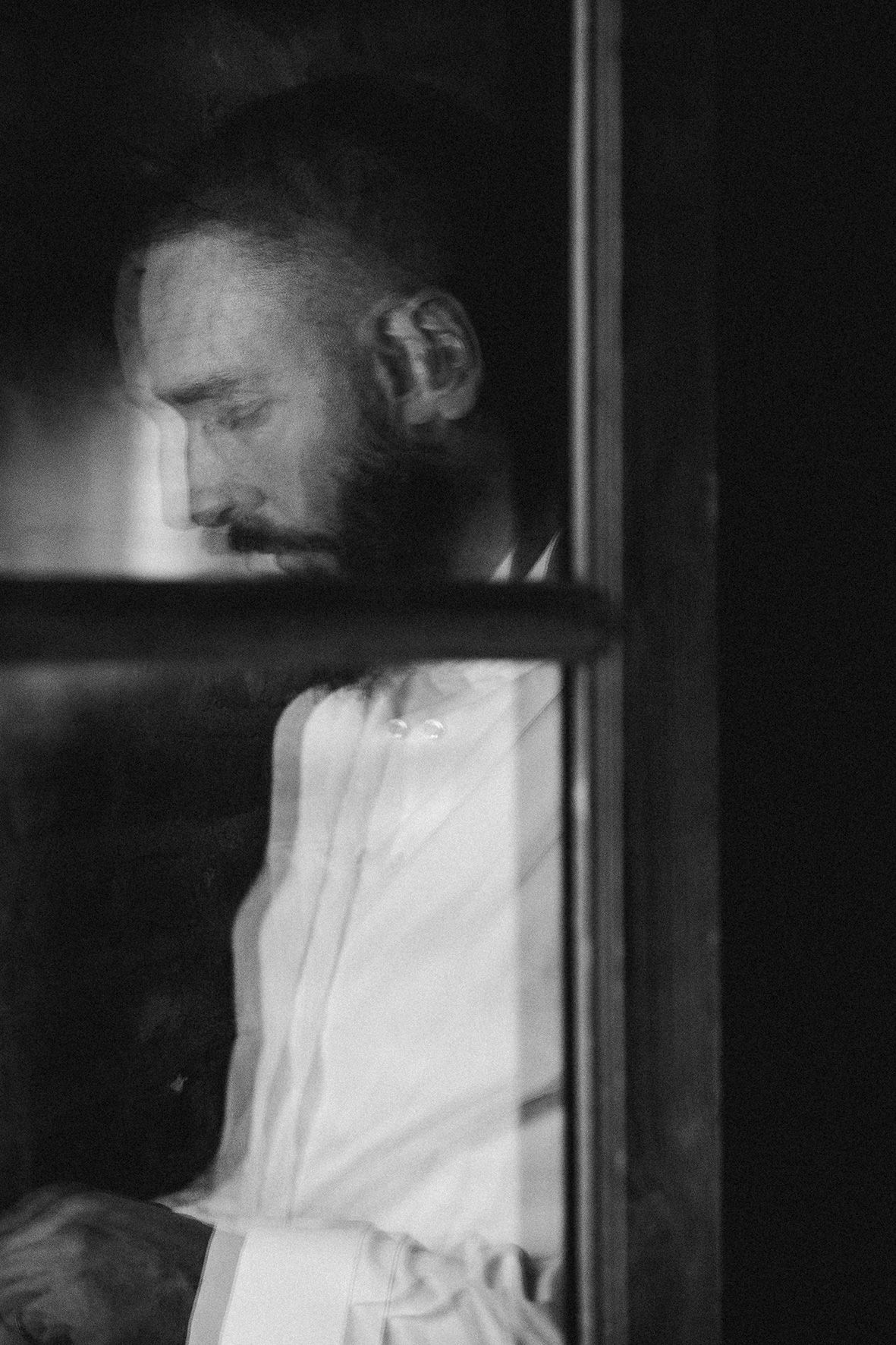 Black and white profile view through a window showing a bearded figure in white clothing.
