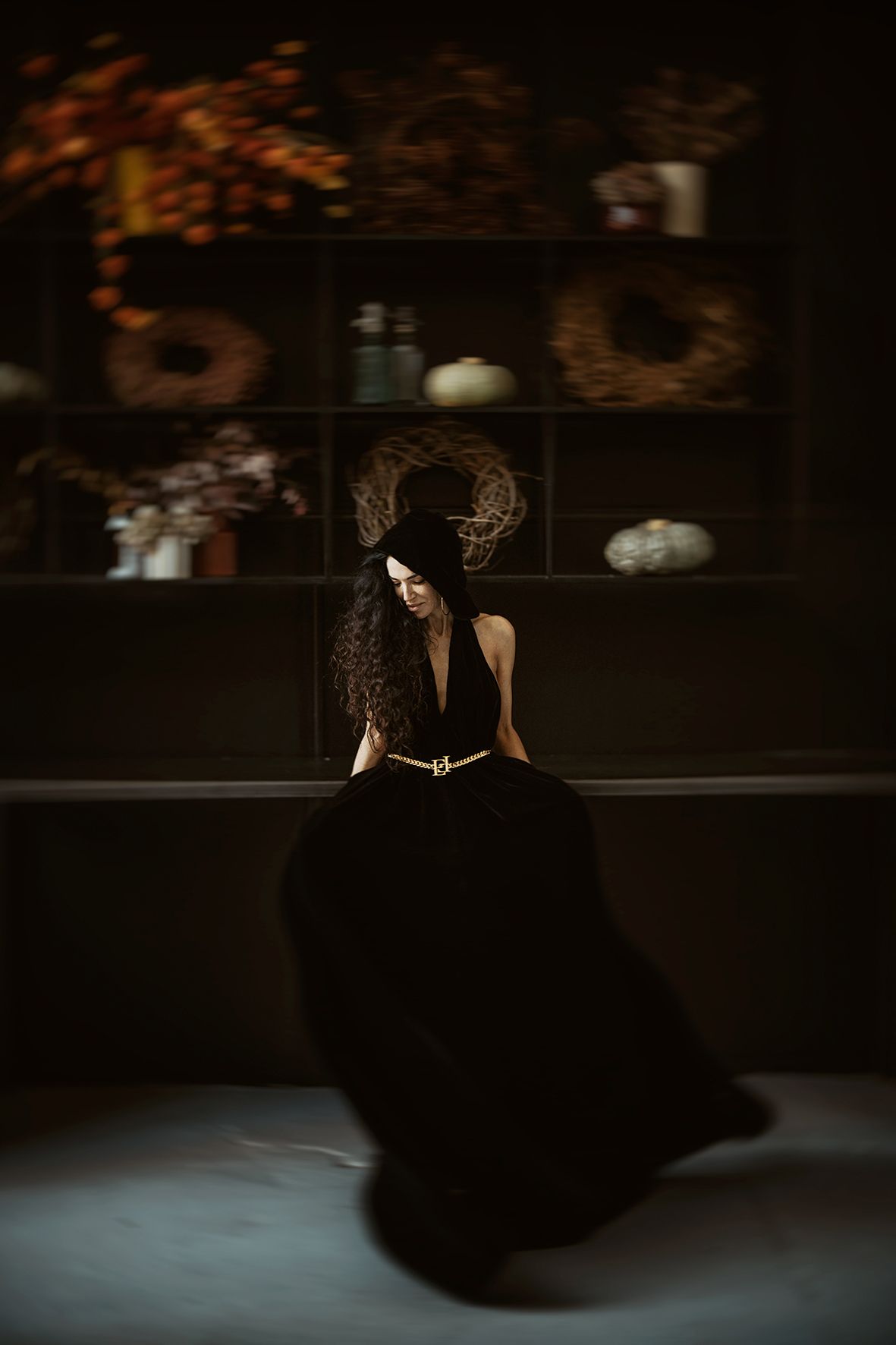 A dramatic indoor portrait featuring a figure in an elegant black ballgown against a dark moody background with decorative shelves.