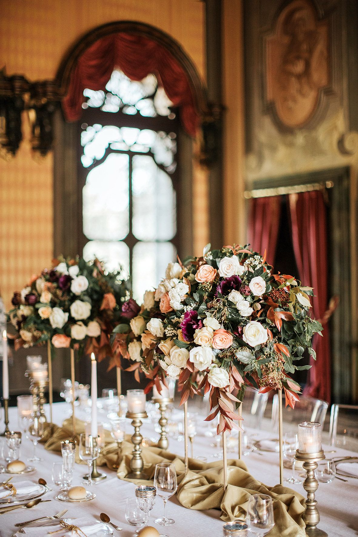 Elegant wedding reception table with lush floral centerpieces in peach and burgundy, crystal candelabras, and gothic windows.