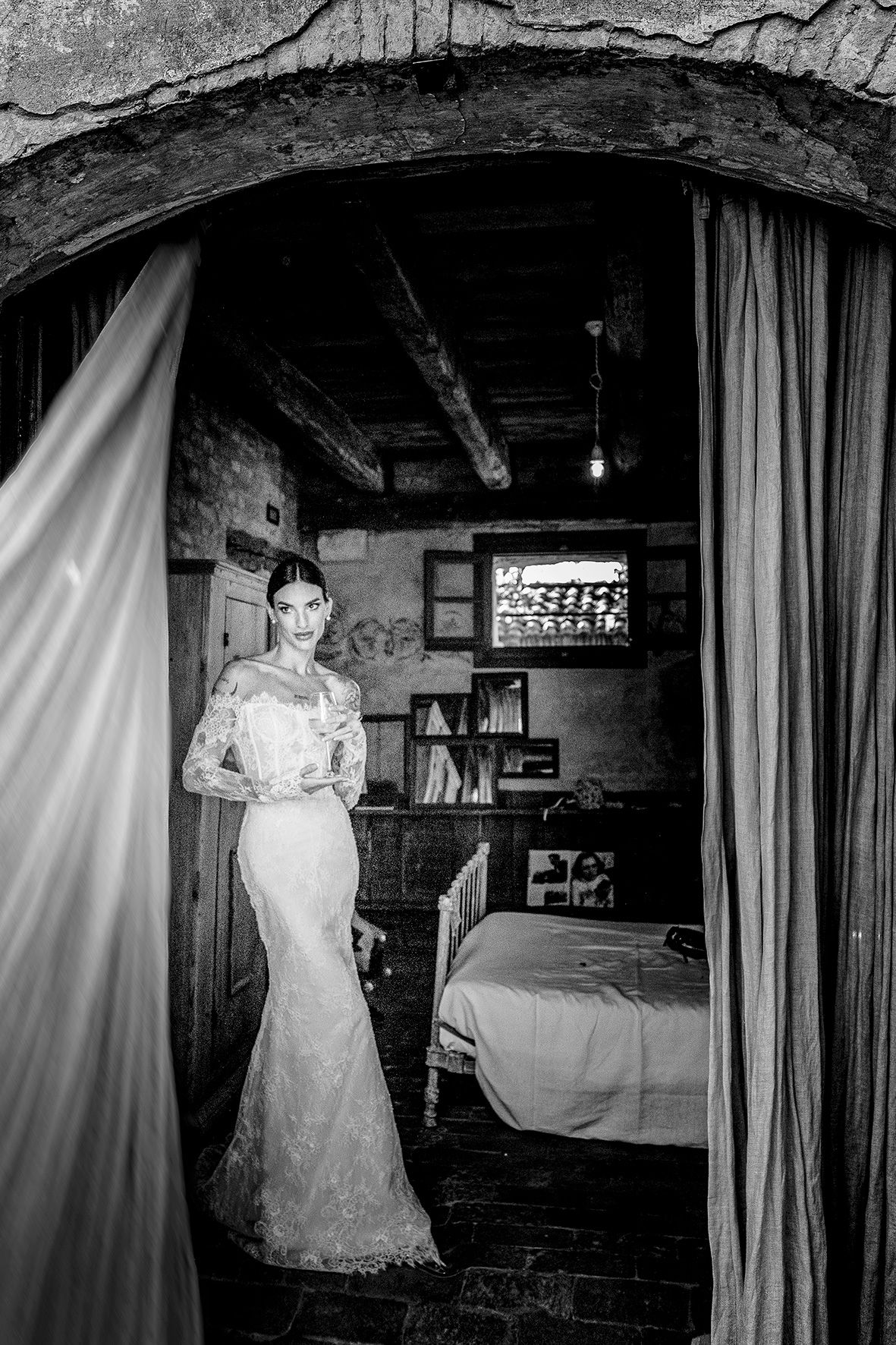 Black and white photo showing someone in an elegant lace wedding dress standing in a rustic room with draped curtains.