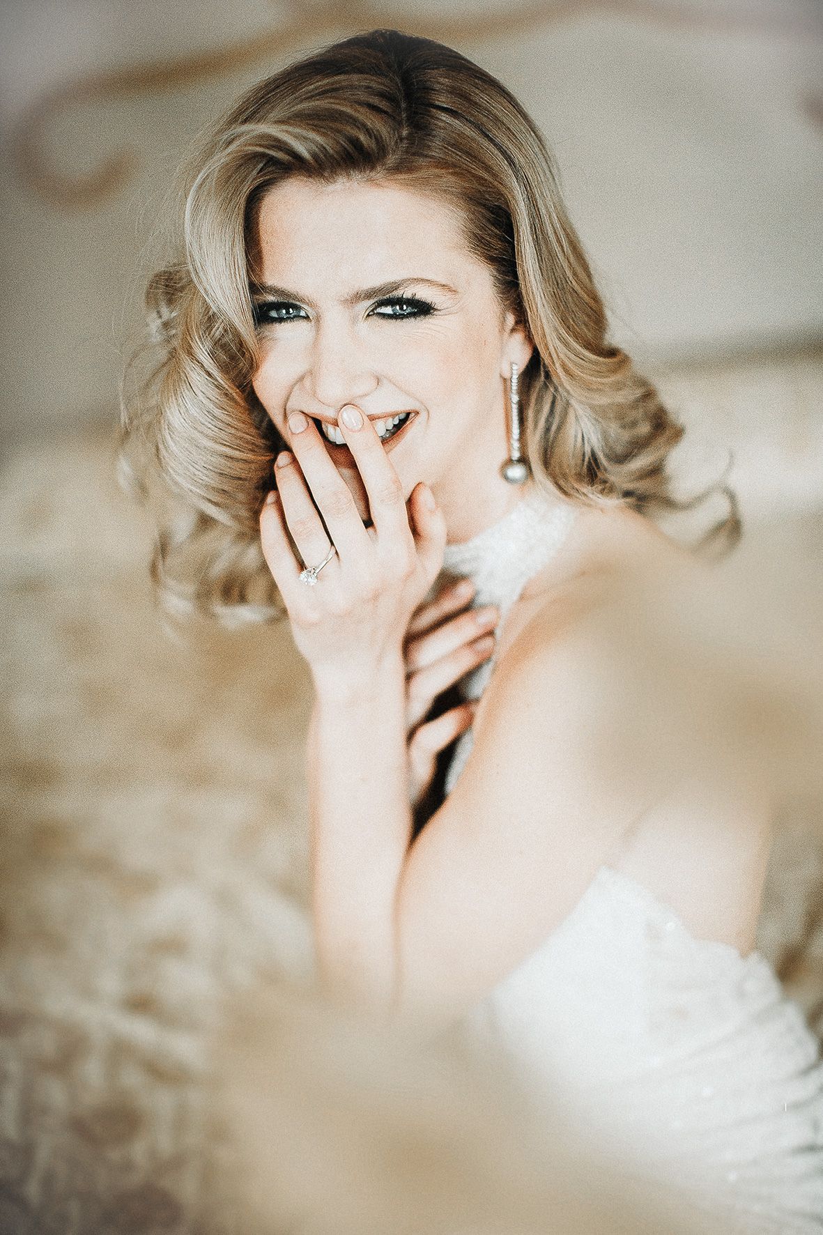 Joyful portrait of a laughing person in a white dress with blonde wavy hair and elegant makeup covering mouth with hand.