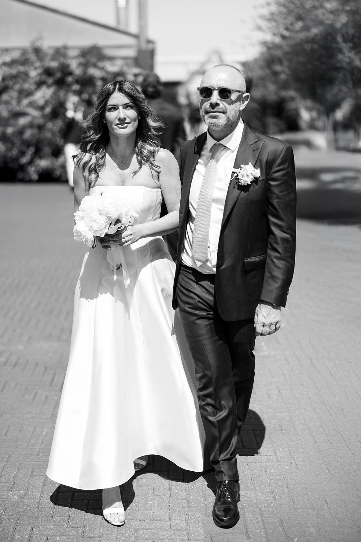 Black and white photo of couple walking outdoors on wedding day with bride holding bouquet.