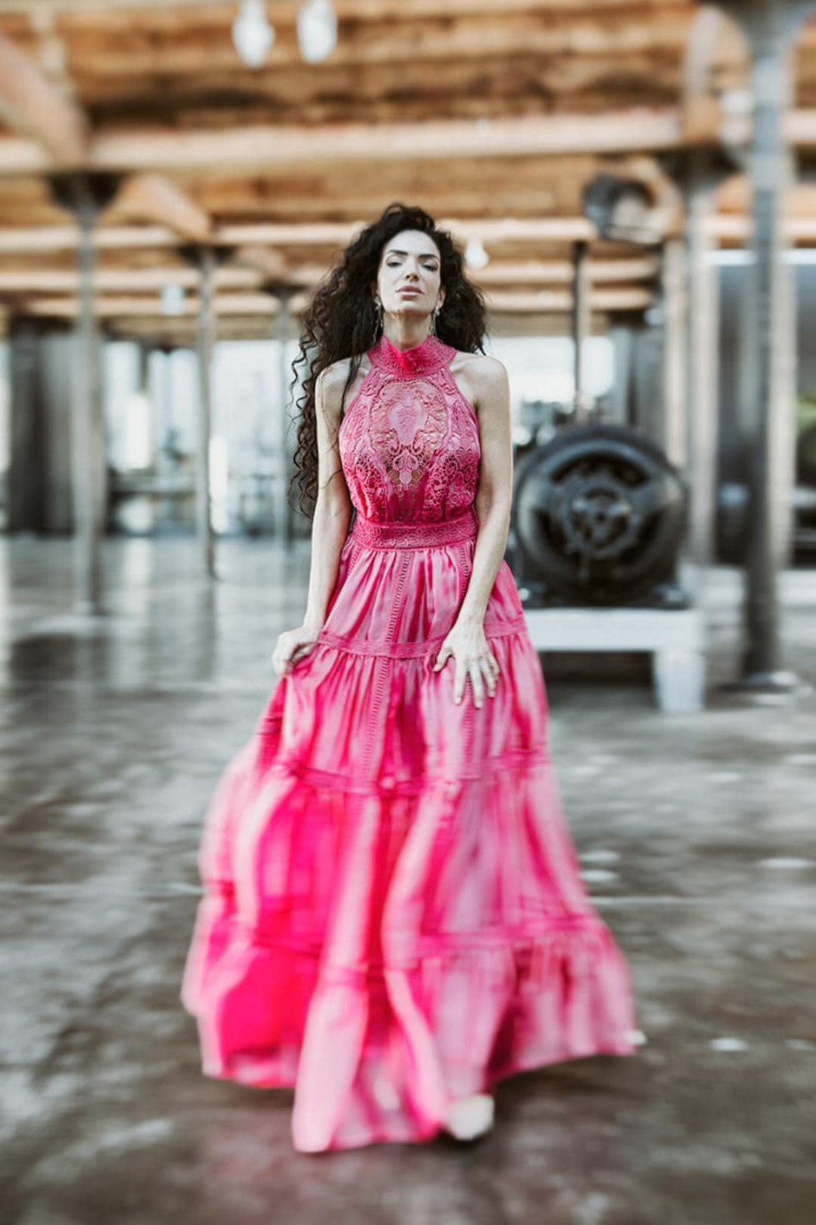 A woman wearing a flowing pink halter gown poses in an industrial setting with rustic wooden beams overhead.