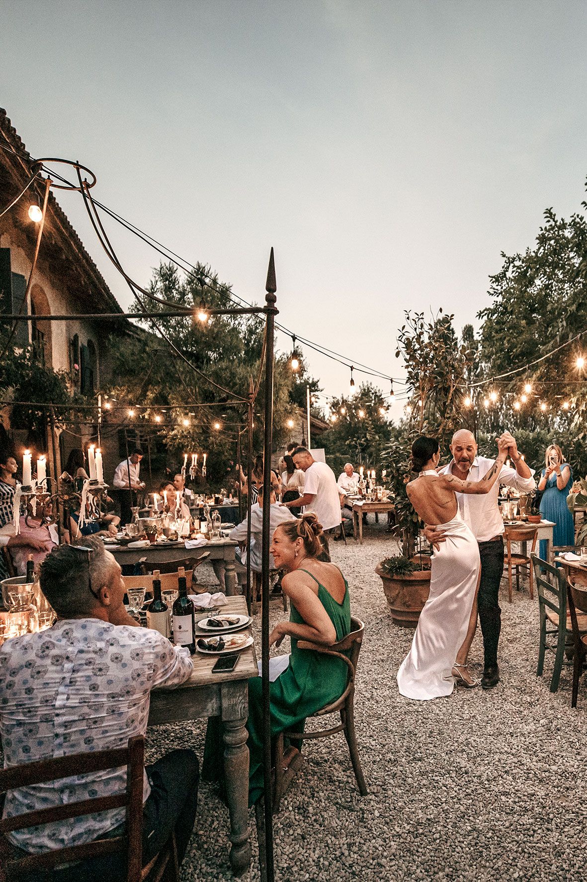Romantic outdoor wedding reception with string lights, rustic tables and guests dining and dancing at dusk.