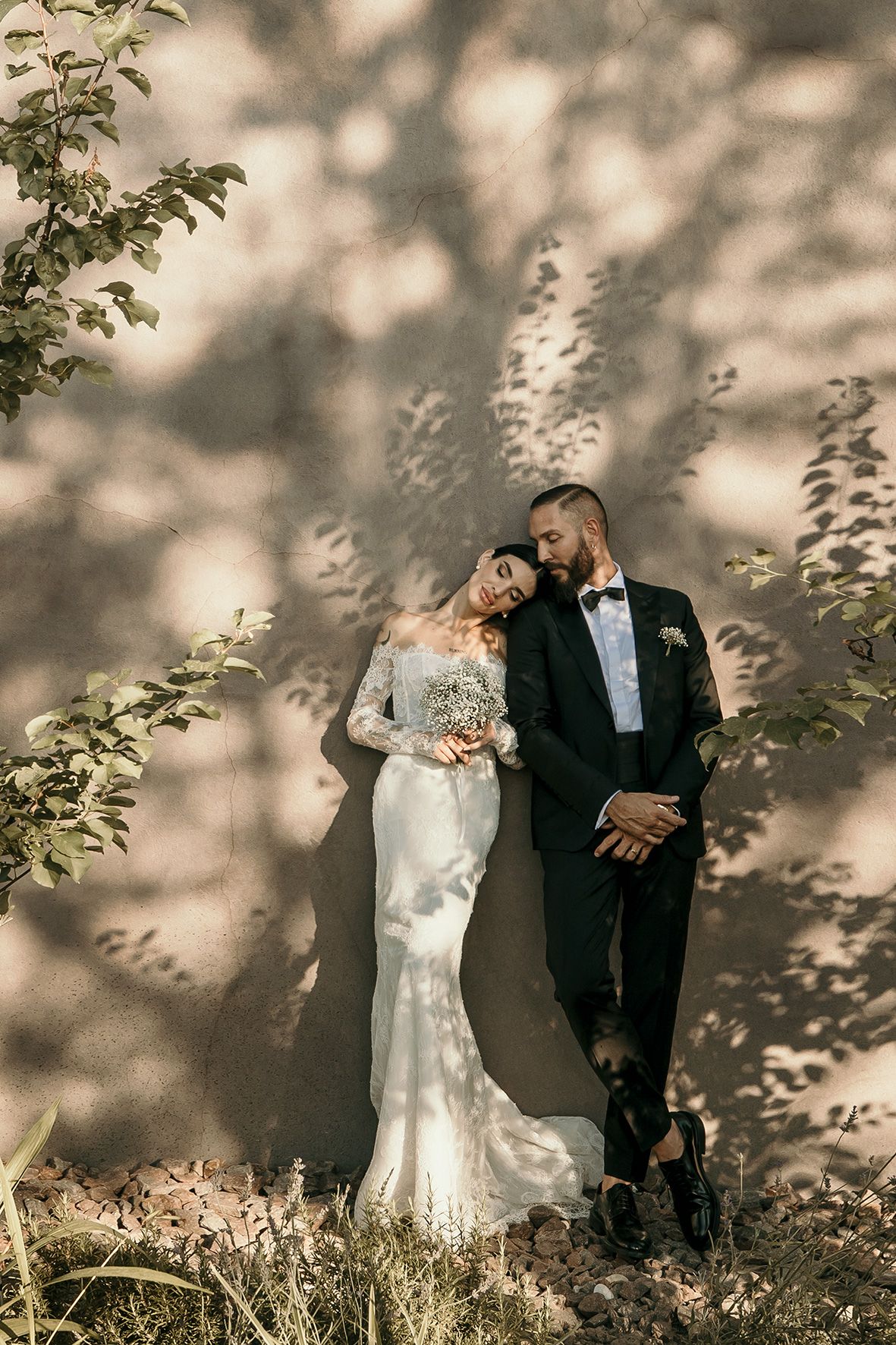 A romantic wedding couple stands together against a wall with dramatic dappled sunlight and tree shadows.