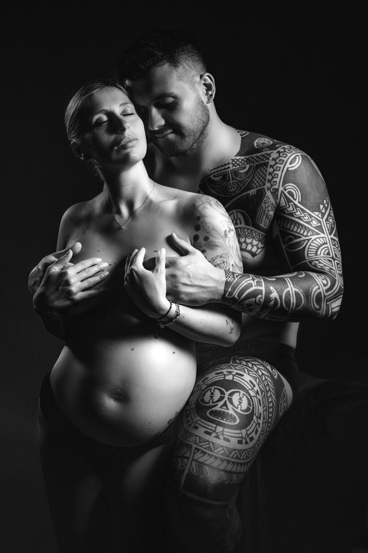 Black and white intimate maternity photo showing a pregnant woman being embraced by partner with tribal tattoos.