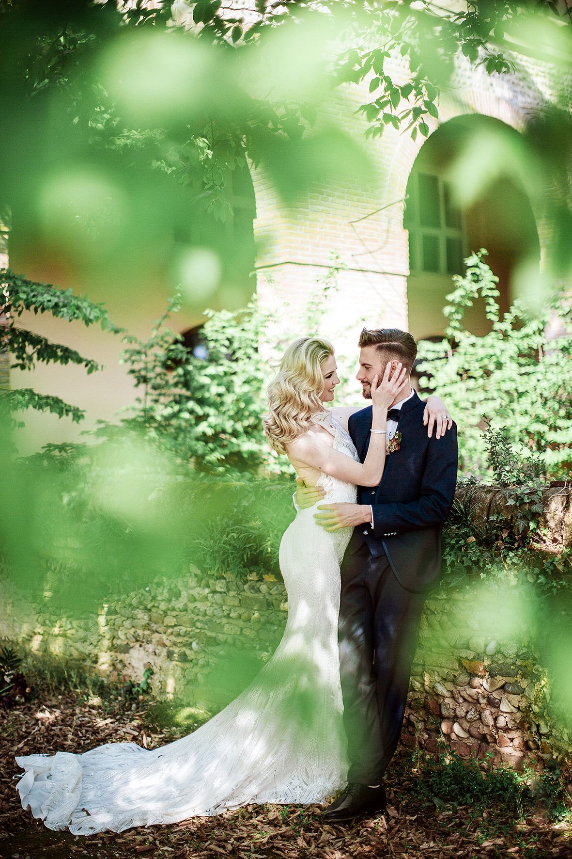A romantic wedding portrait framed through blurred greenery and foliage in a garden setting.