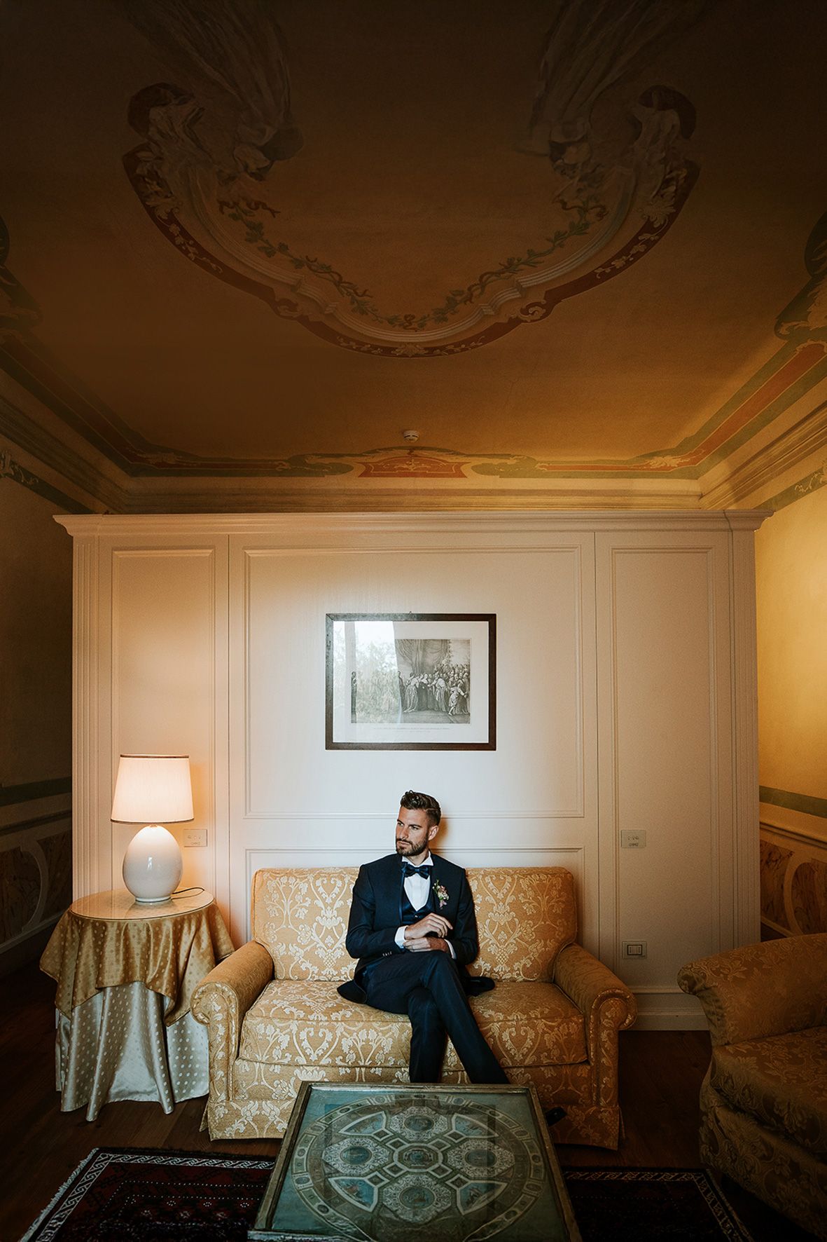 Groom in classic tuxedo sits on vintage sofa in elegant room with ornate ceiling details and warm lighting.