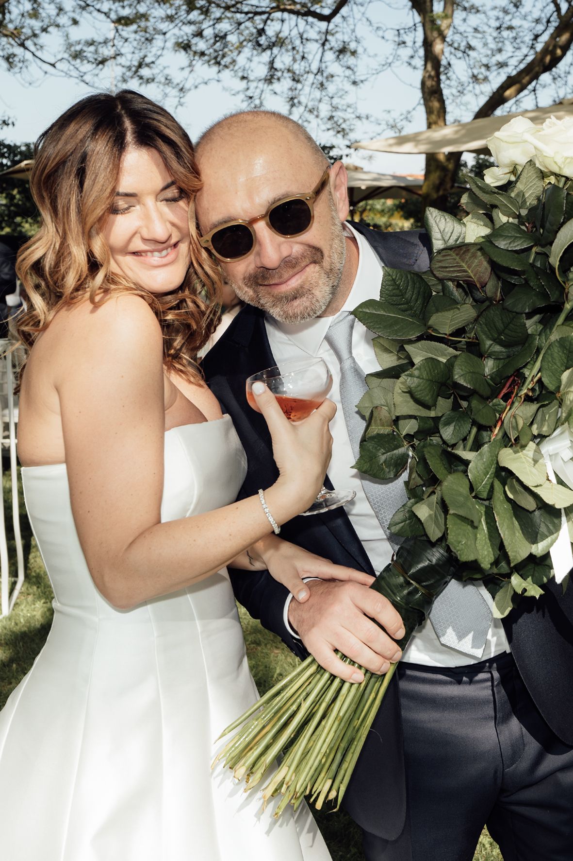 Newlywed couple at outdoor wedding celebration holding wine glasses and bouquet with greenery backdrop.