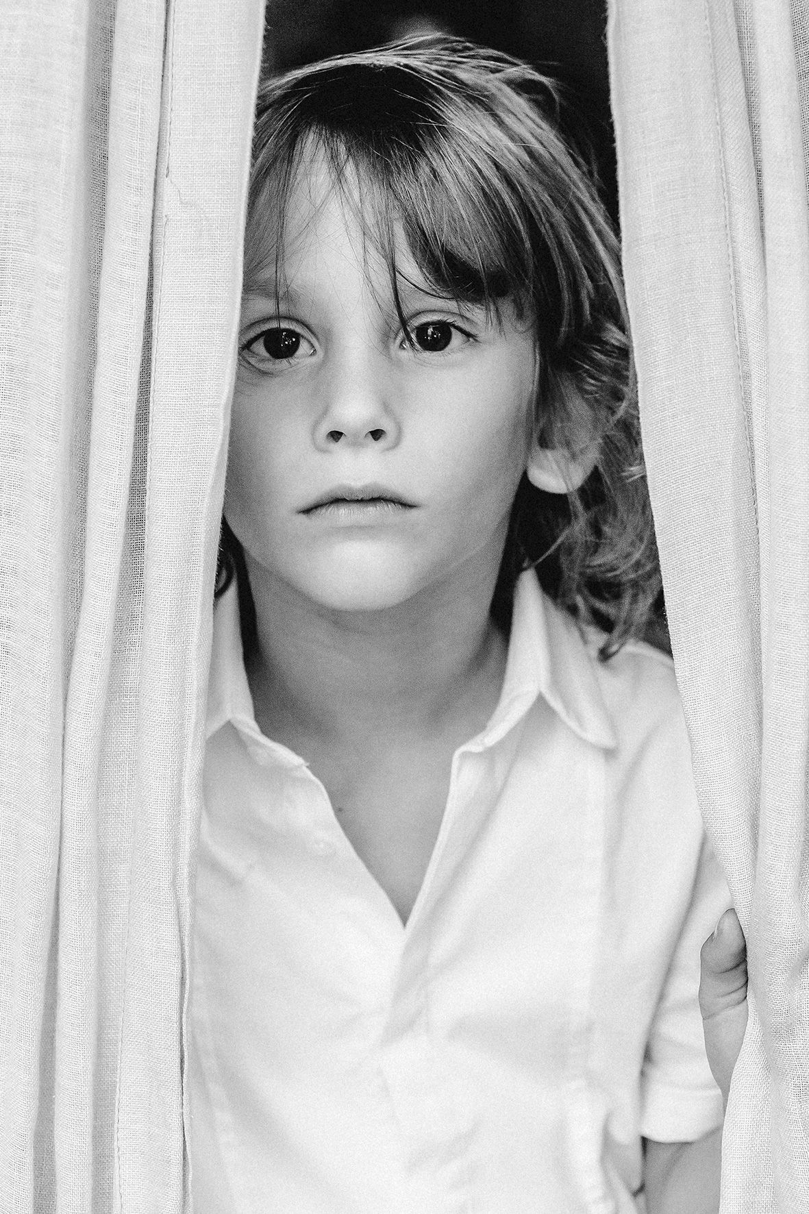 Black and white portrait of a young child in a white shirt looking intently at camera against light curtain background.