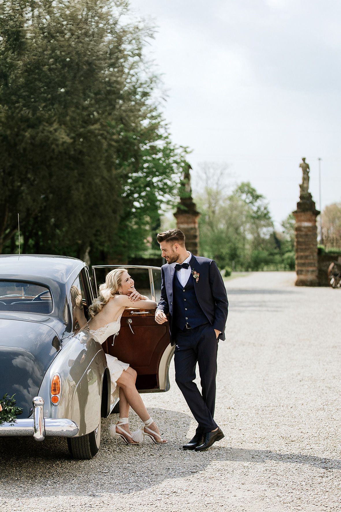 Couple poses by vintage car at entrance of elegant driveway with ornate stone pillars and lush trees.