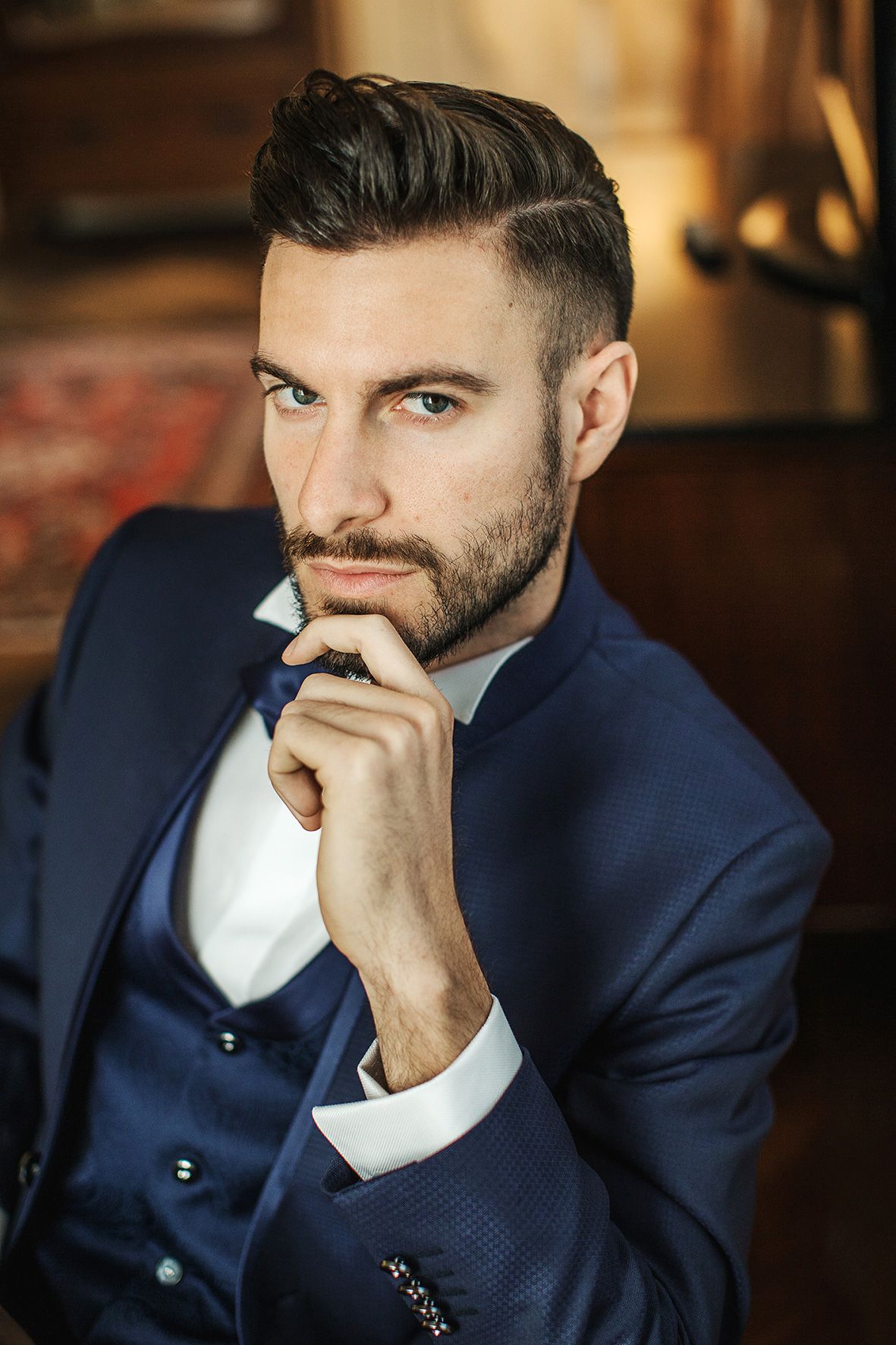 A well-dressed person in a tailored navy blue suit and white shirt poses thoughtfully in an elegant setting.