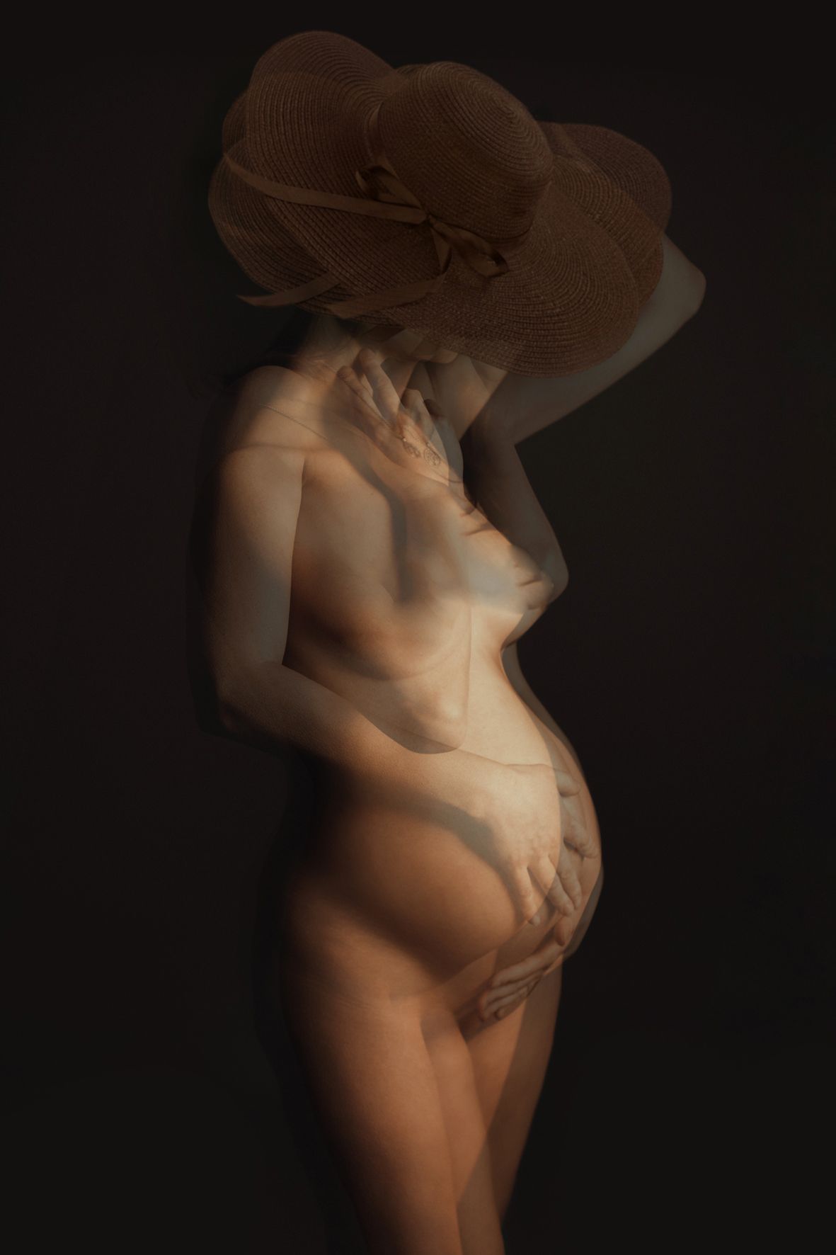 Artistic nude maternity silhouette against dark background featuring a brown sunhat and soft lighting.