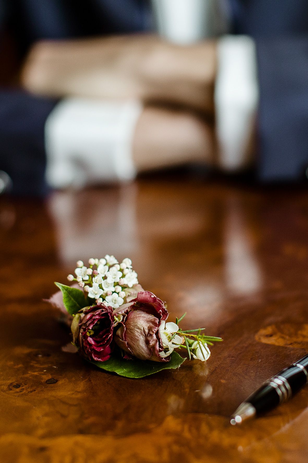 A delicate boutonniere with burgundy roses and baby's breath rests on a polished wooden surface next to a pen.