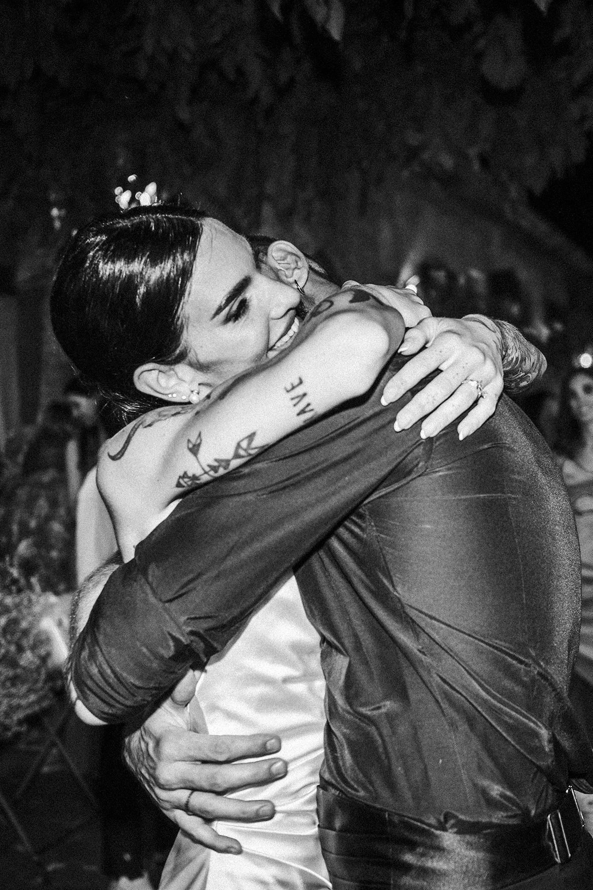 Black and white photo of two people sharing an intimate embrace at what appears to be a wedding celebration.