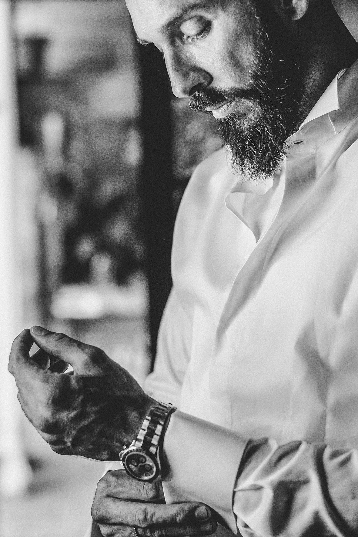 Black and white close-up of a bearded person wearing a white shirt adjusting their wristwatch.