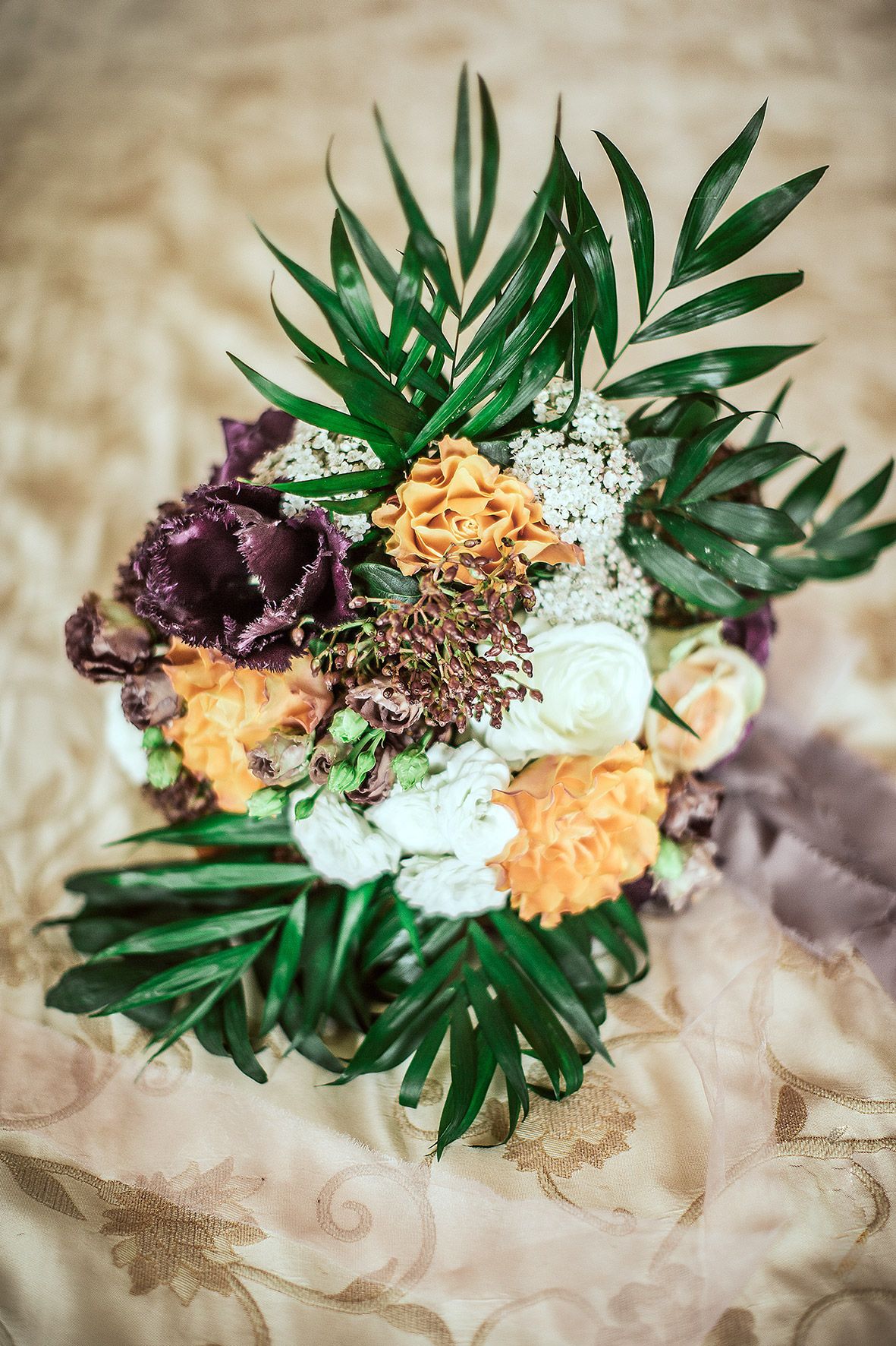 A wedding bouquet with purple and peach flowers surrounded by green palm leaves rests on light fabric.