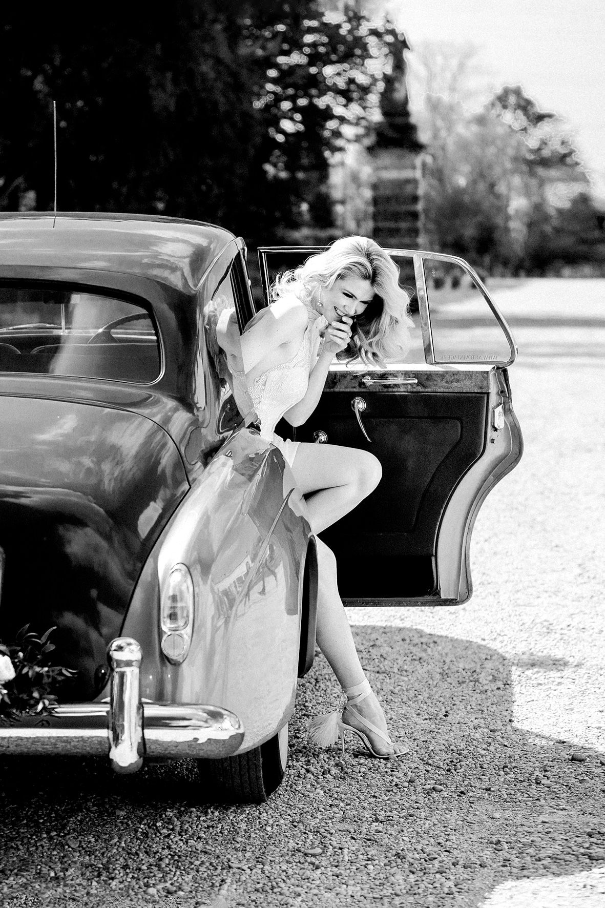 A vintage black and white photograph showing elegant styling with a classic car and heels on a gravel path.