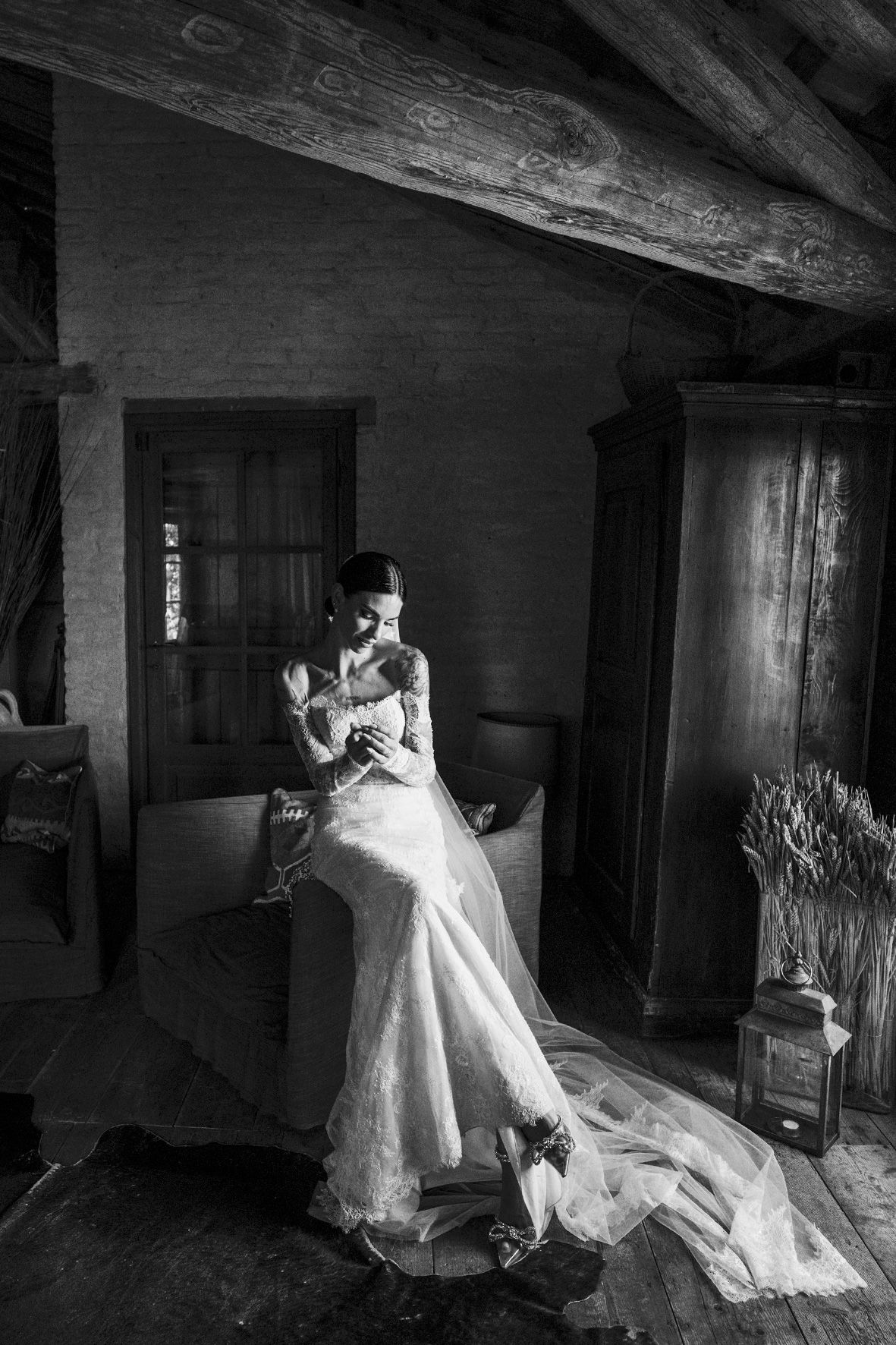 Black and white photograph of a figure in a flowing lace wedding dress sitting in a rustic room with wooden beams.
