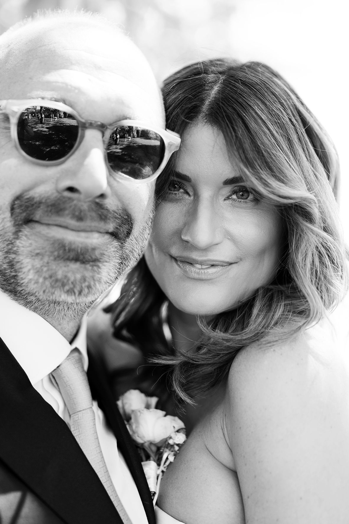 Black and white close-up portrait of a couple at their wedding, with the groom wearing sunglasses and a boutonniere.