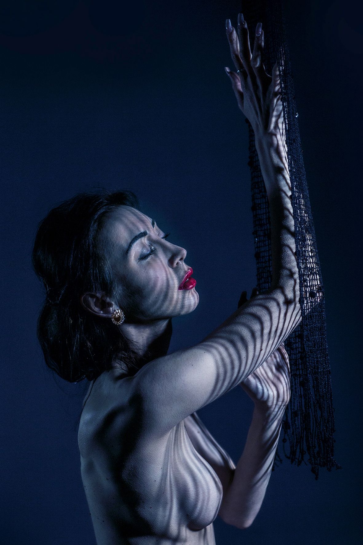 Artistic portrait of someone against blue backdrop holding a decorative black fringe tassel while in dramatic pose.