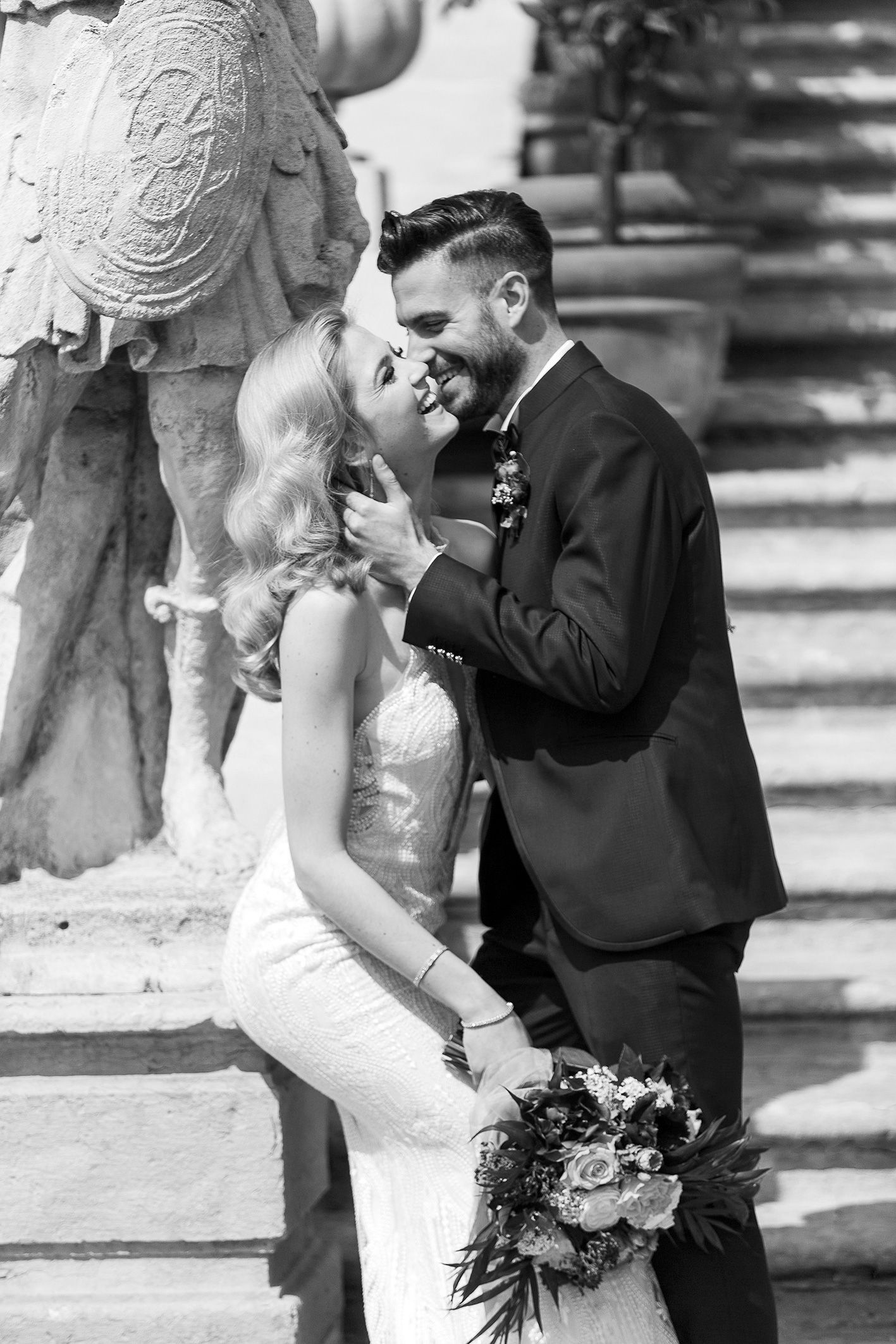 A romantic black and white wedding photo captures a tender moment on stone steps with a floral bouquet.