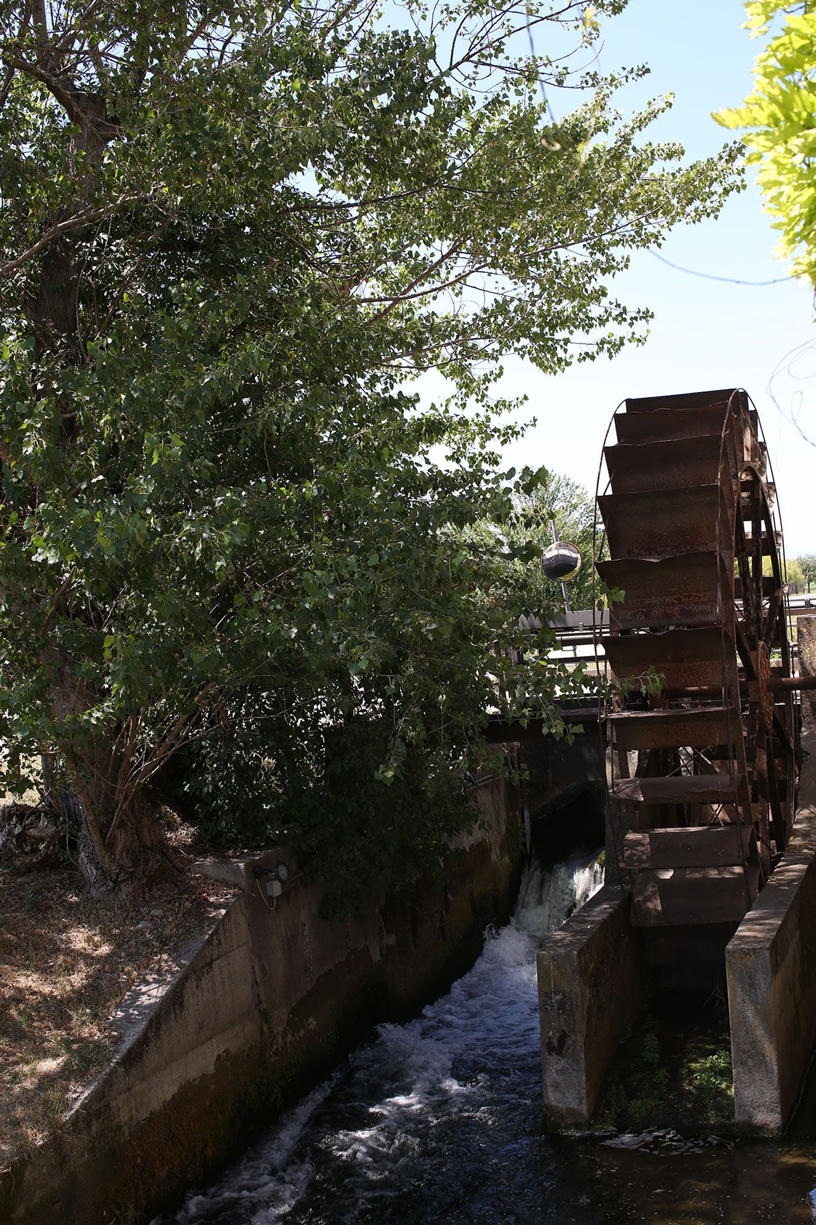 A rustic wooden water wheel stands beside a flowing canal with overhanging trees creating a peaceful rural scene.