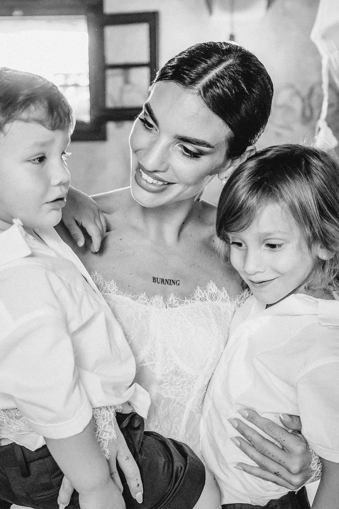 A joyful bride in a white wedding dress shares an intimate moment with two young children in matching white shirts.