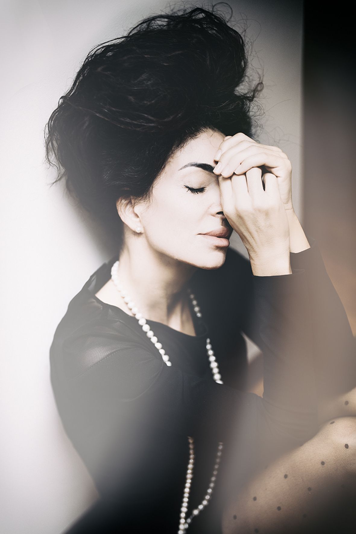 Artistic portrait photograph in black and white featuring dark hair and pearl necklace against soft dreamy background.