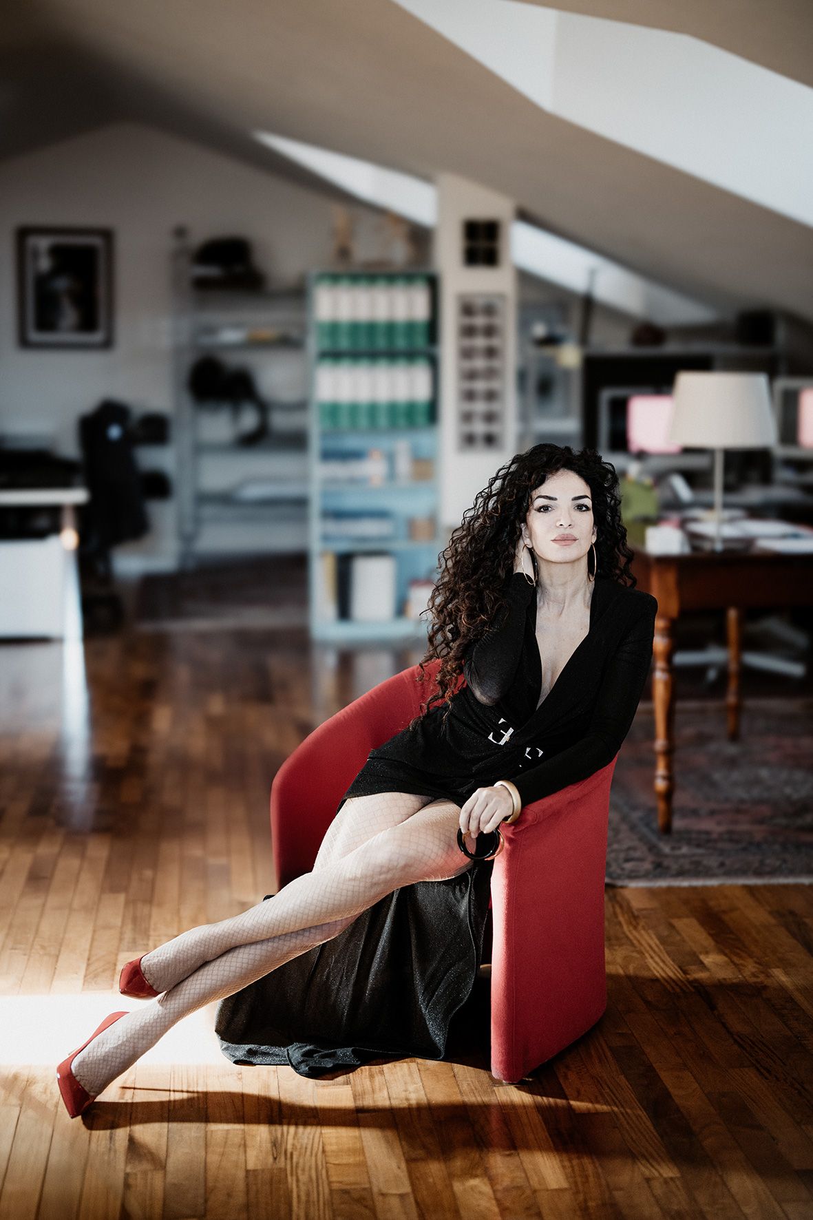 Woman in black dress lounges elegantly on red chair in modern loft space with hardwood floors.