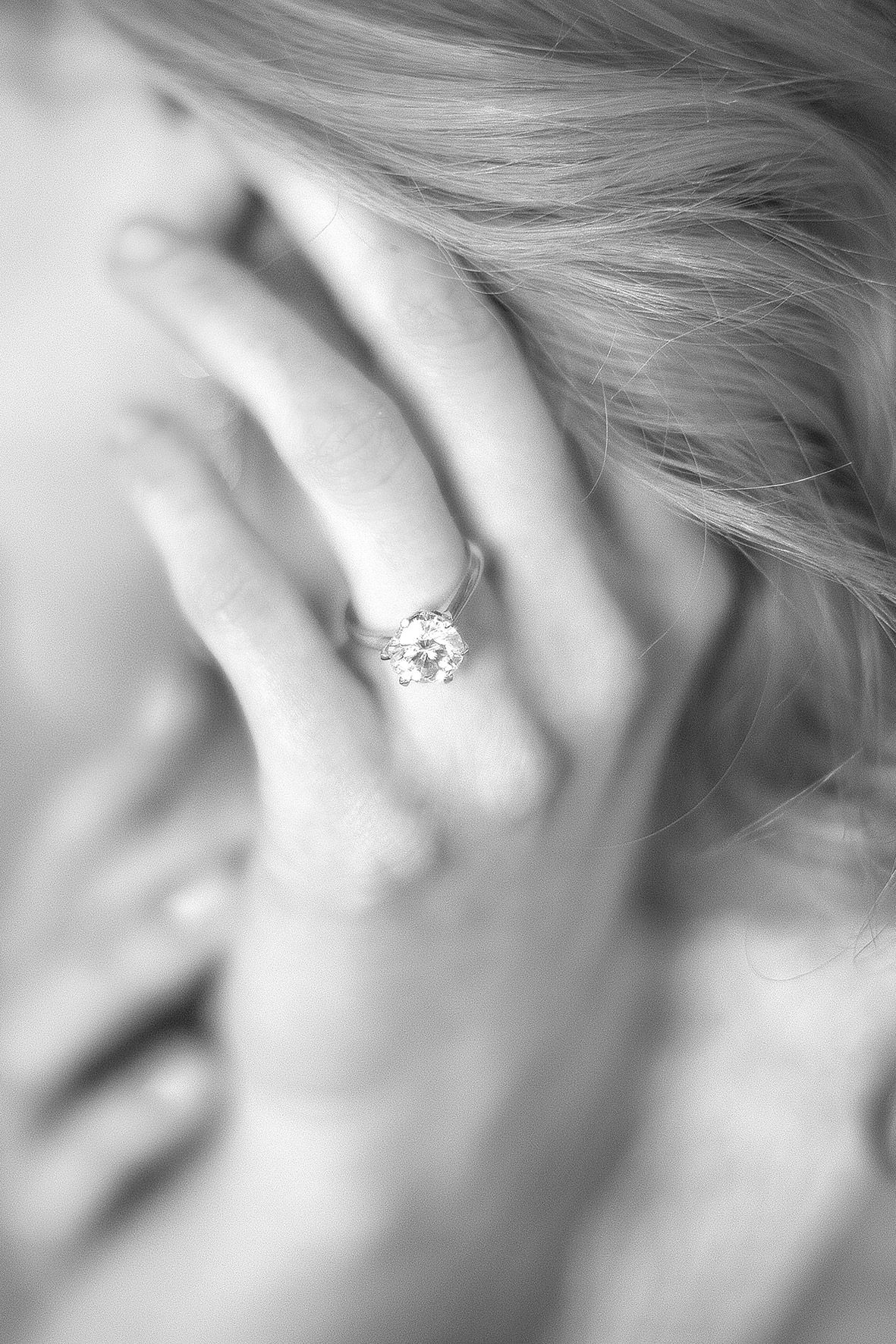 A close-up black and white photo shows a sparkling diamond engagement ring worn on a delicate hand.