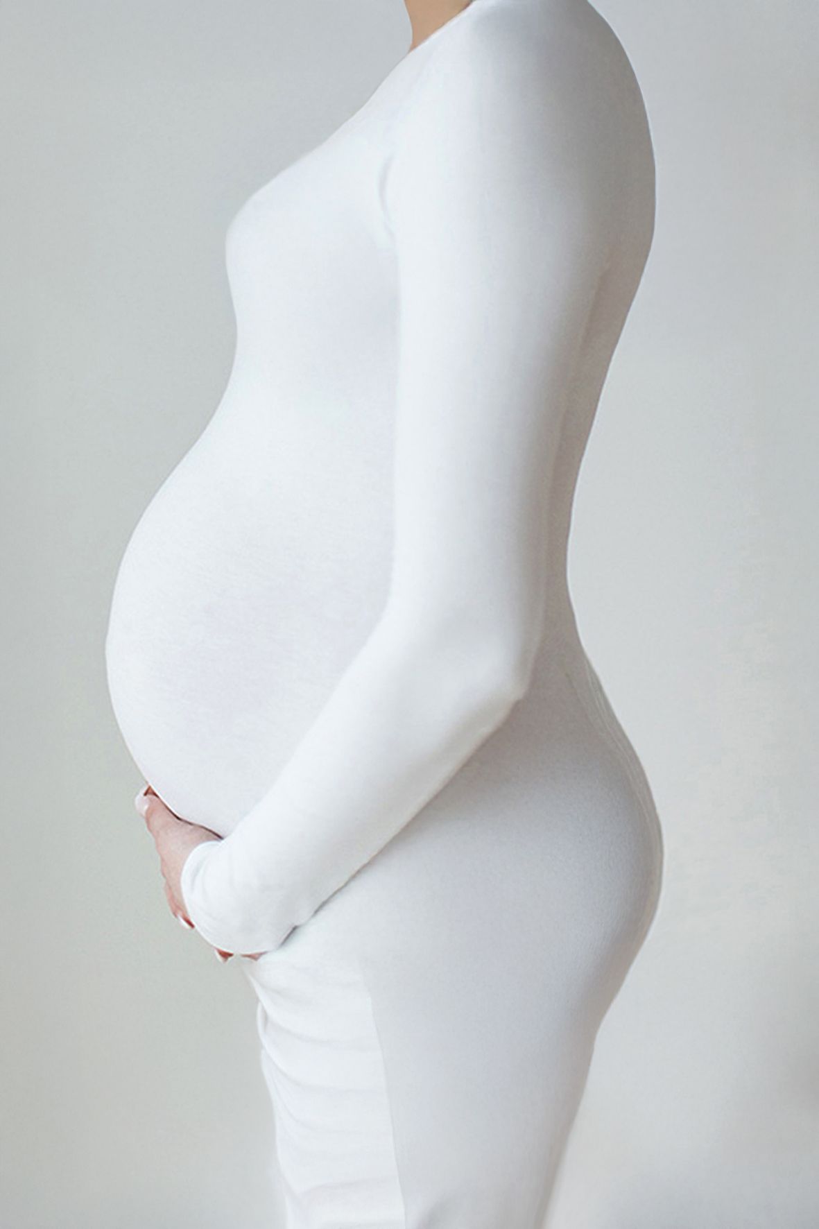 Side profile view of a pregnant woman wearing a form-fitting white dress showing her baby bump against a light background.