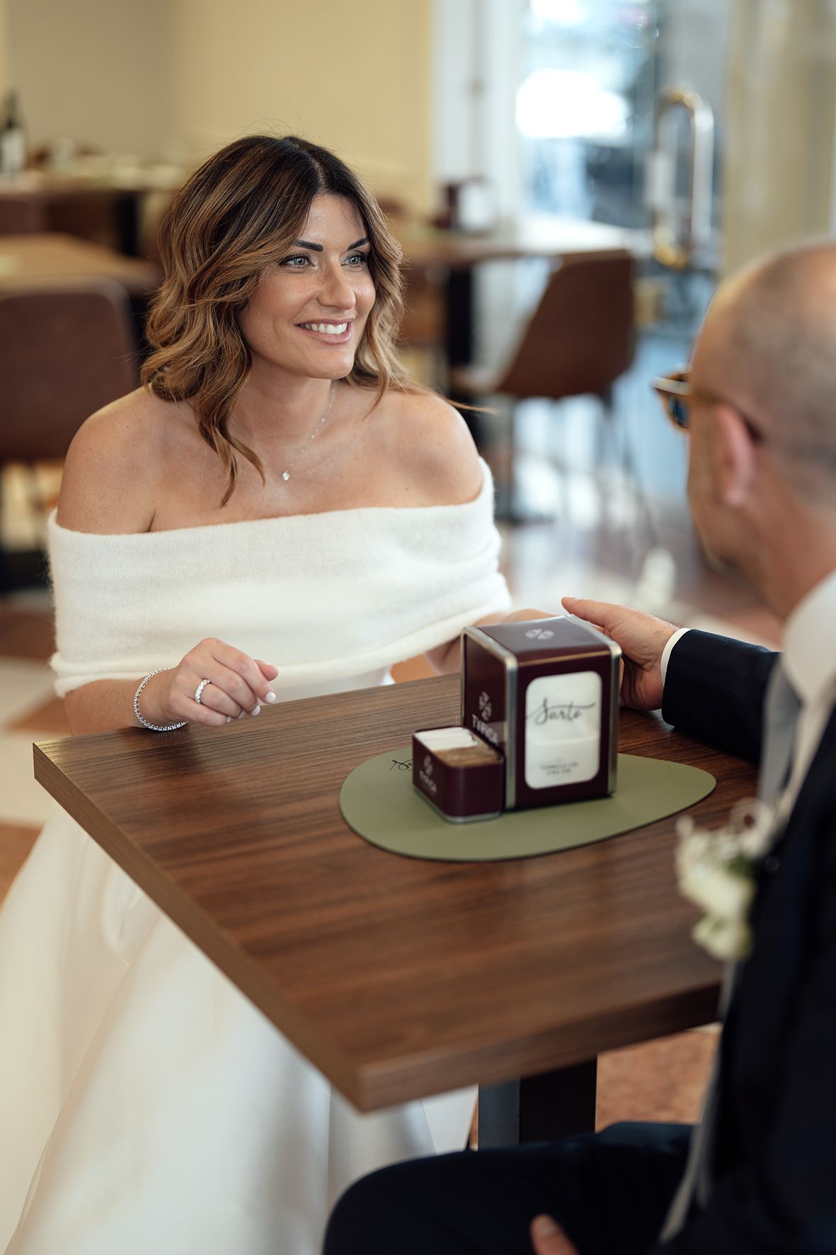 An intimate moment at a restaurant table shows a person in a white off-shoulder dress looking at jewelry boxes with their companion.