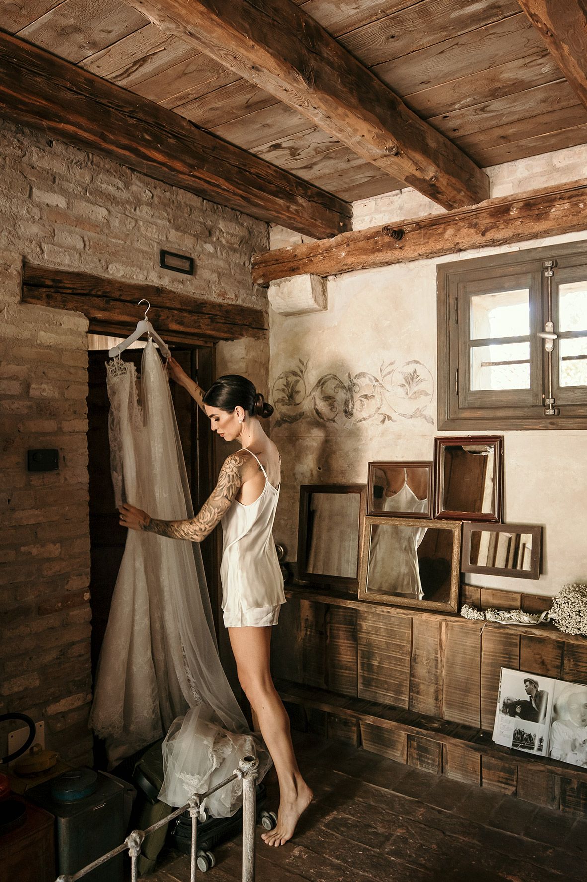 A wedding dress hangs in a rustic room with exposed wooden beams, stone walls and vintage frames leaning against the wall.