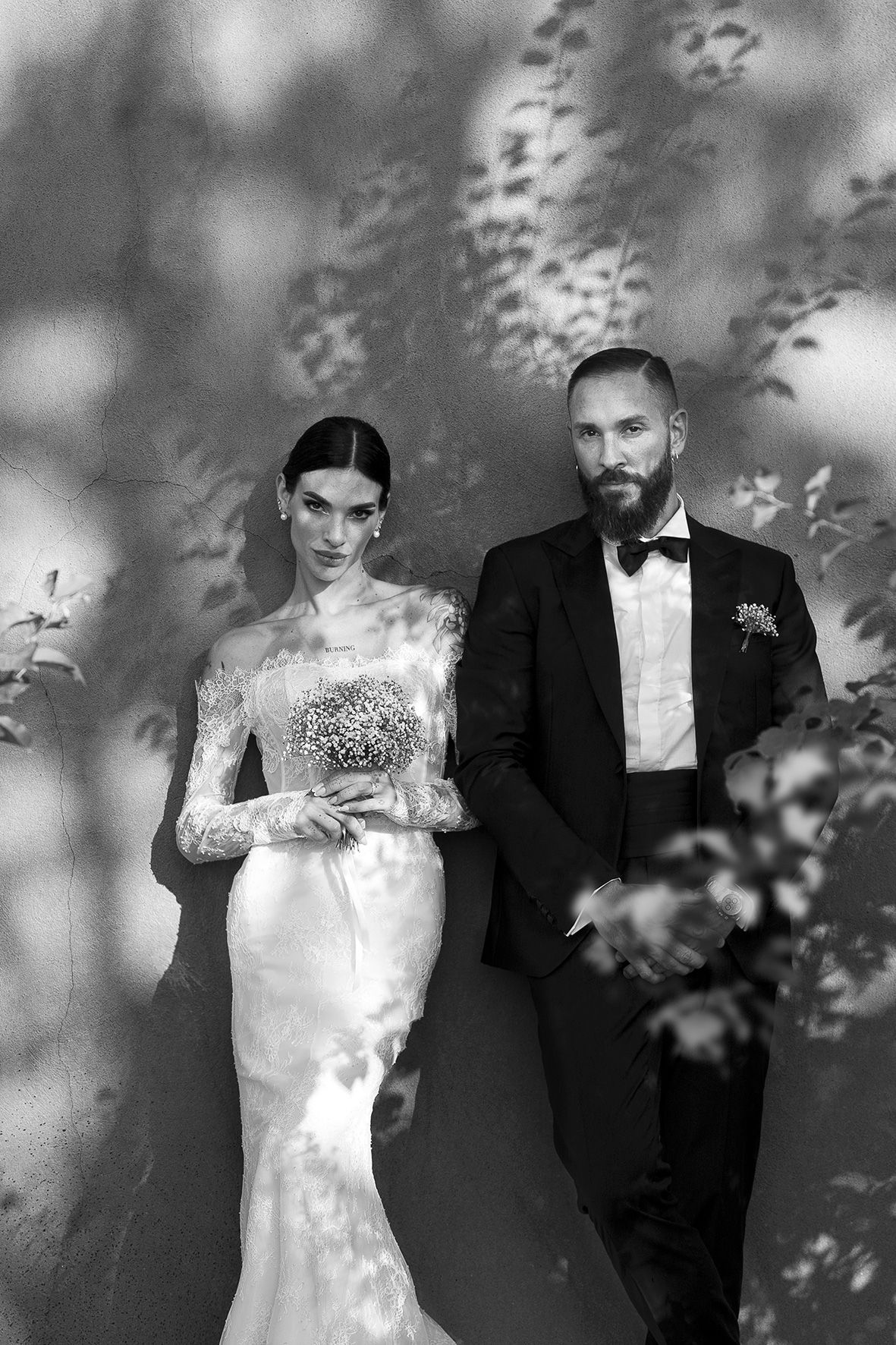 Black and white wedding photo of a couple posing together with delicate leaf shadows cast on the wall behind them.