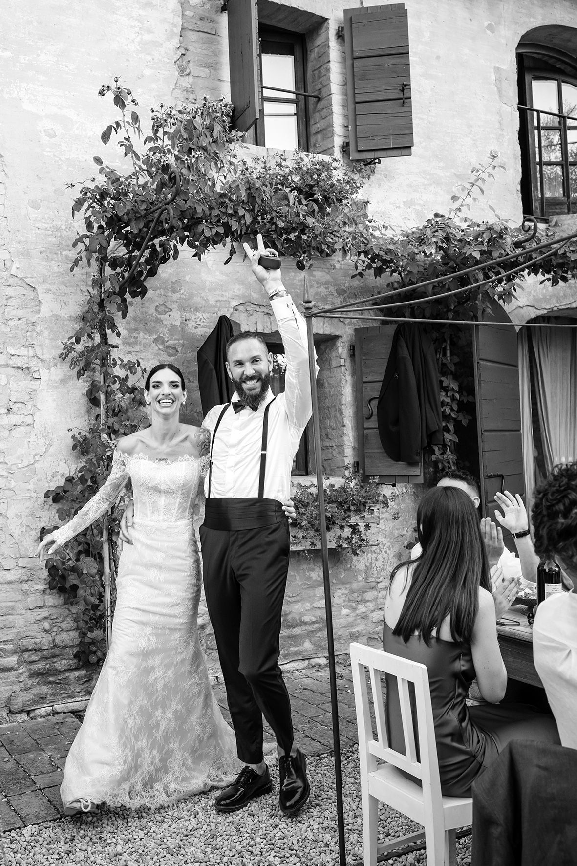 A black and white wedding photo showing a celebratory moment in front of a rustic stone building with climbing vines.
