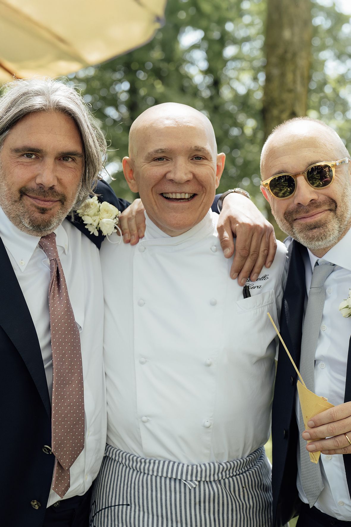 Three well-dressed men pose together outdoors, with the middle person wearing a white chef's uniform and striped pants.