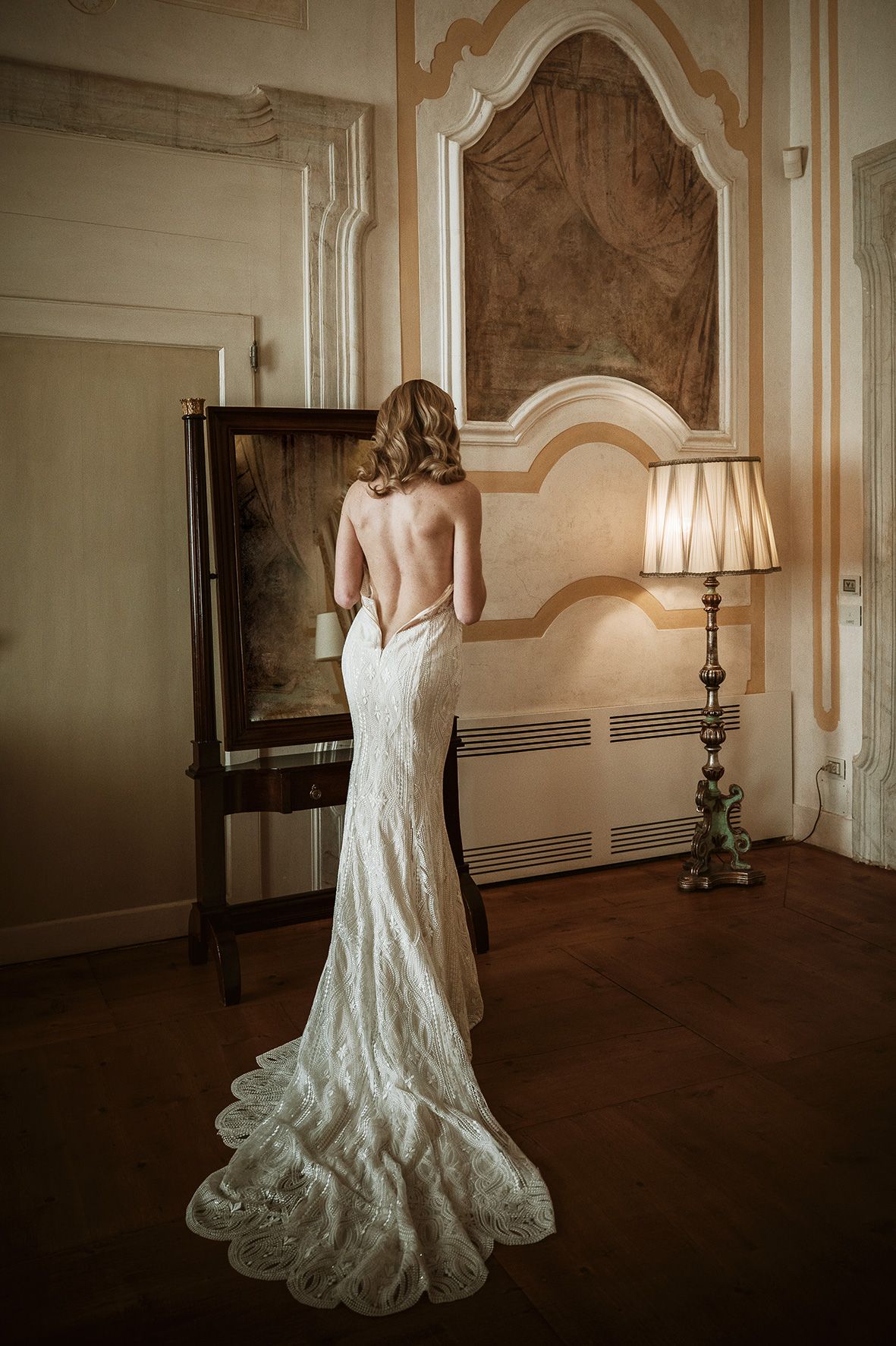 A bride in a backless lace wedding gown with a flowing train stands in an elegant room with antique furniture and decor.