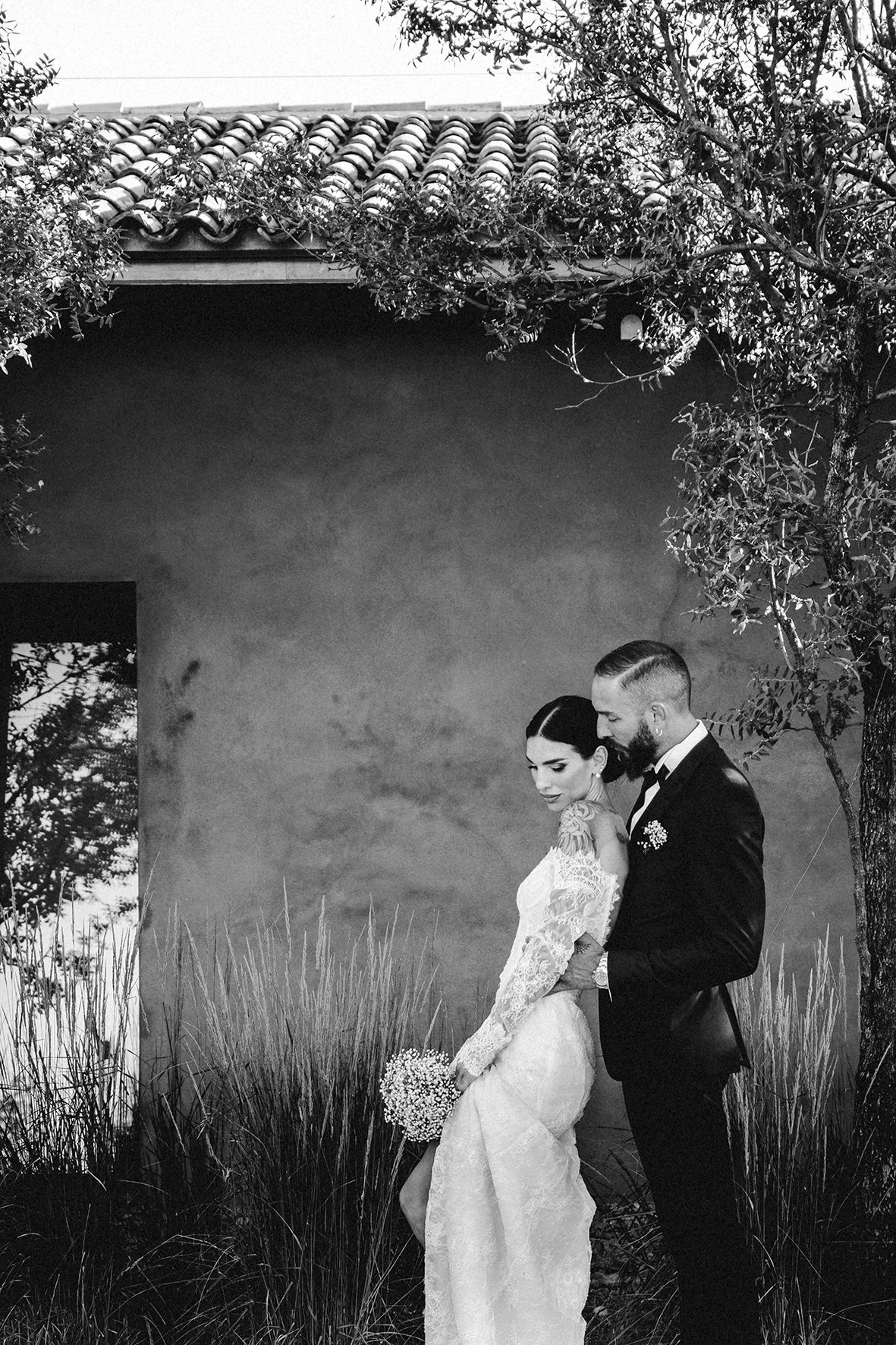 A romantic black and white wedding photo showing a couple posing outdoors against a stucco wall with ornate tile roofing.