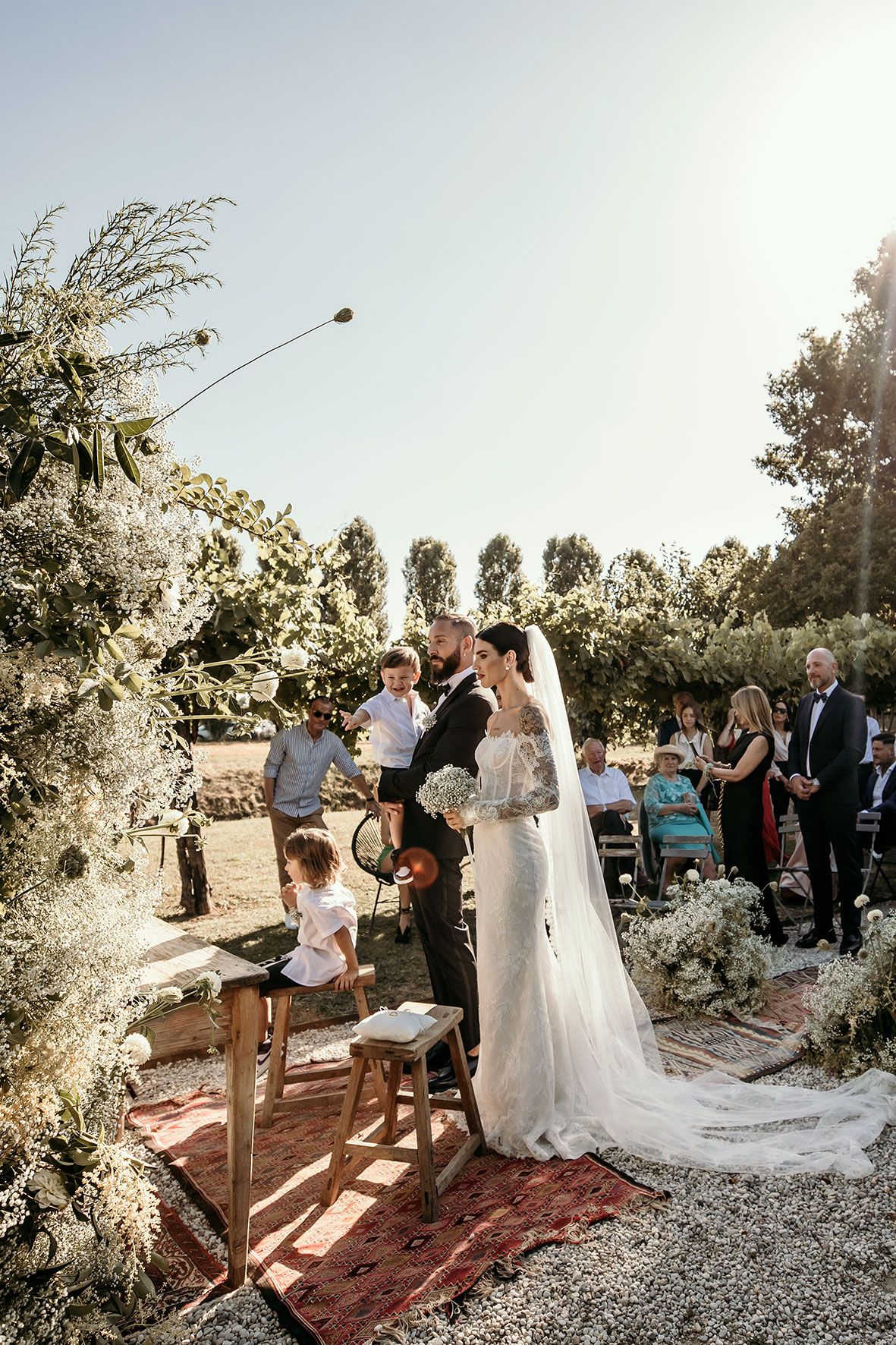 A romantic outdoor wedding ceremony set among sunlit trees with floral arrangements and guests gathered on a gravel path.