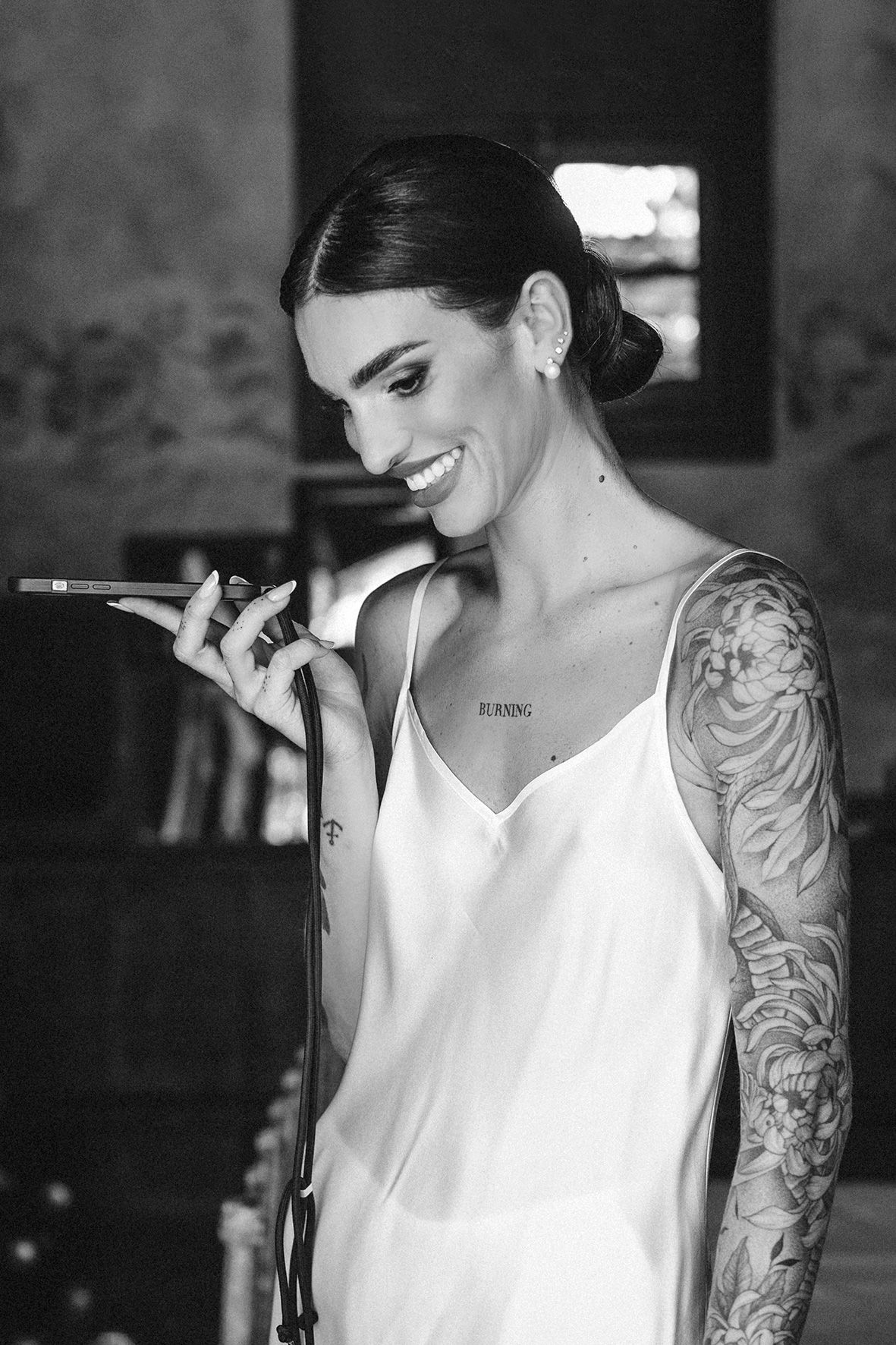 Black and white photo shows person in white top smiling while holding phone, displaying intricate arm tattoo sleeve.