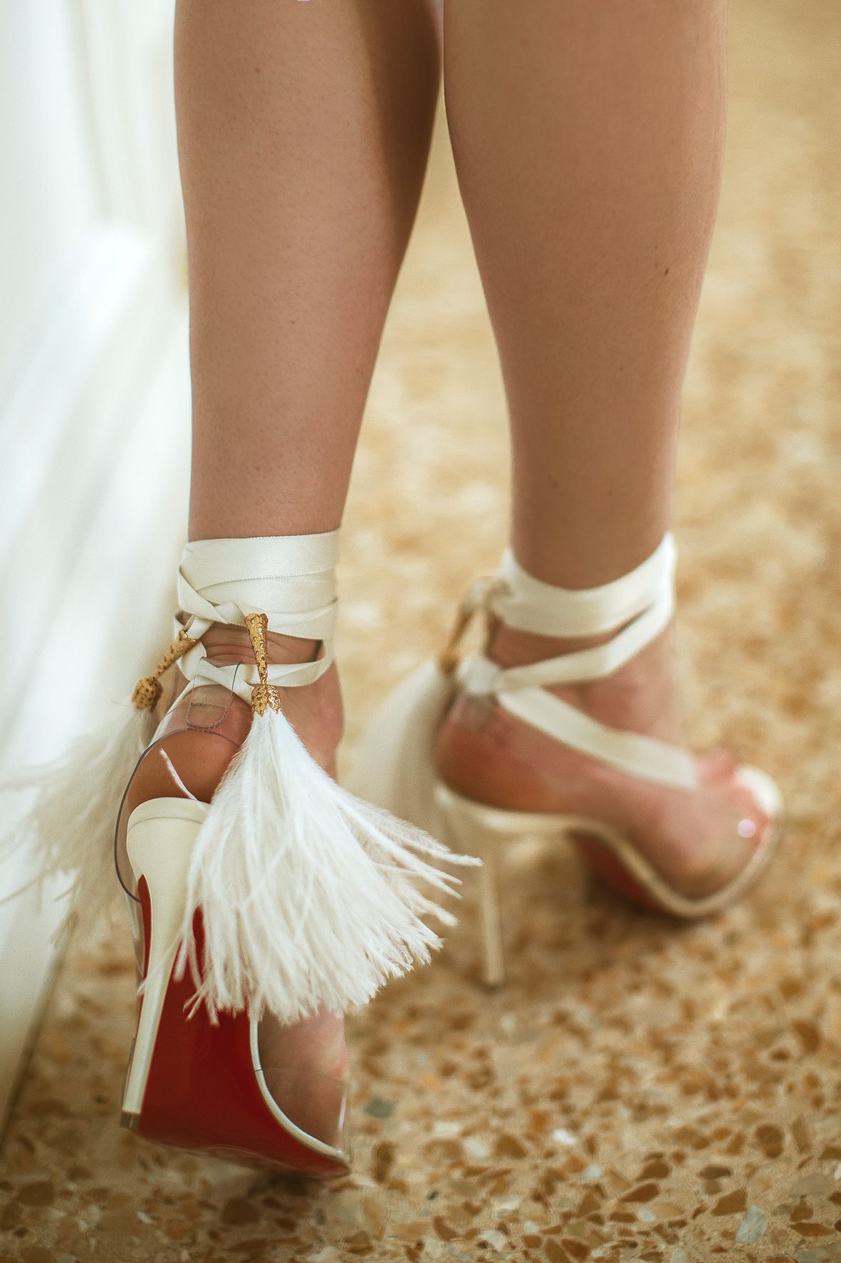 White leather high heels with feather tassels and ankle straps photographed against textured flooring.